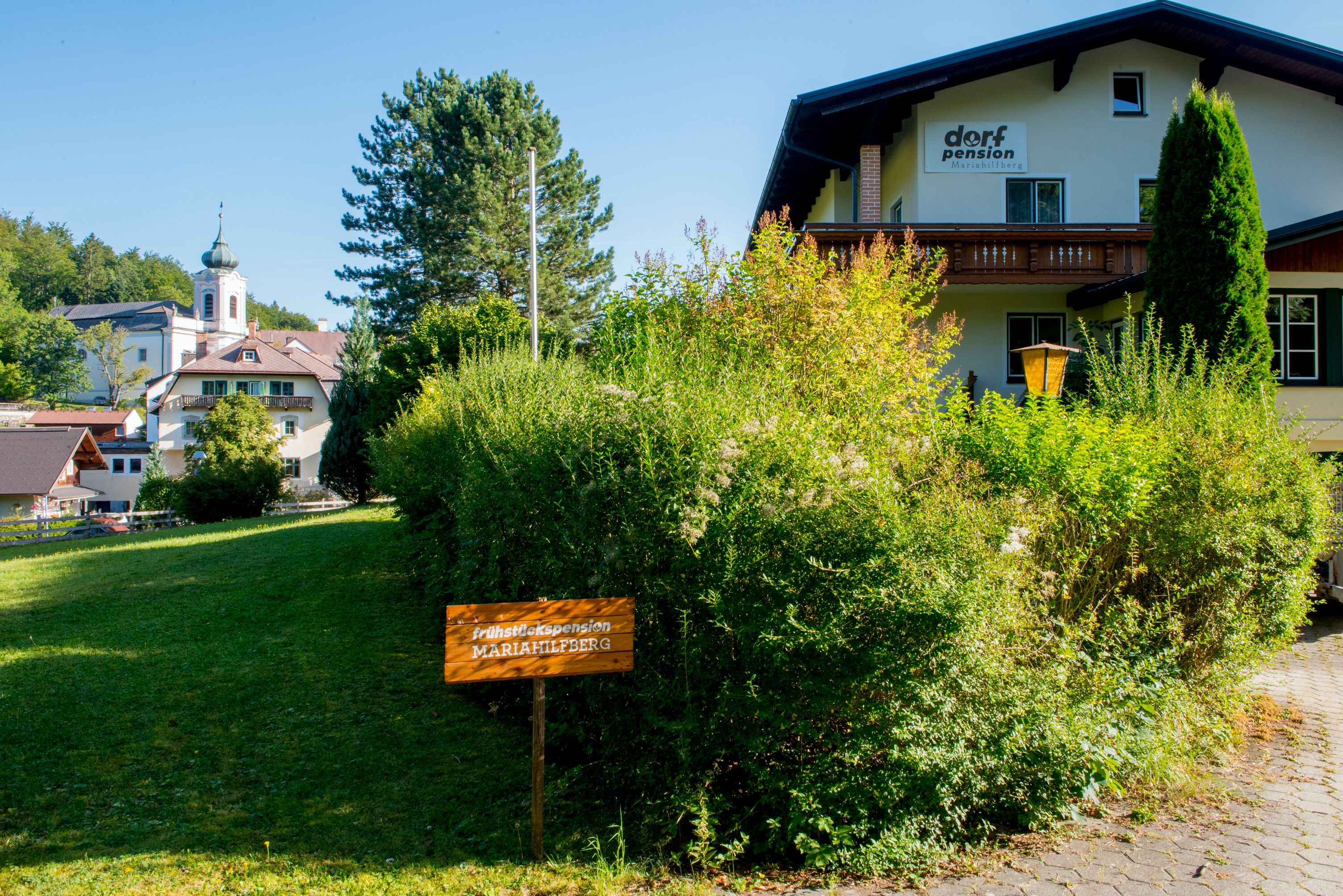 Budova s označením "Dorfpension" a nápisom "Frühstückspension Mariahilfberg" v zelenom prostredí.