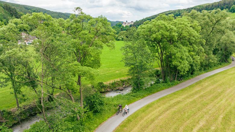 Dvaja cyklisti na ceste pozdĺž rieky obklopenej zelenými stromami a lúkami.