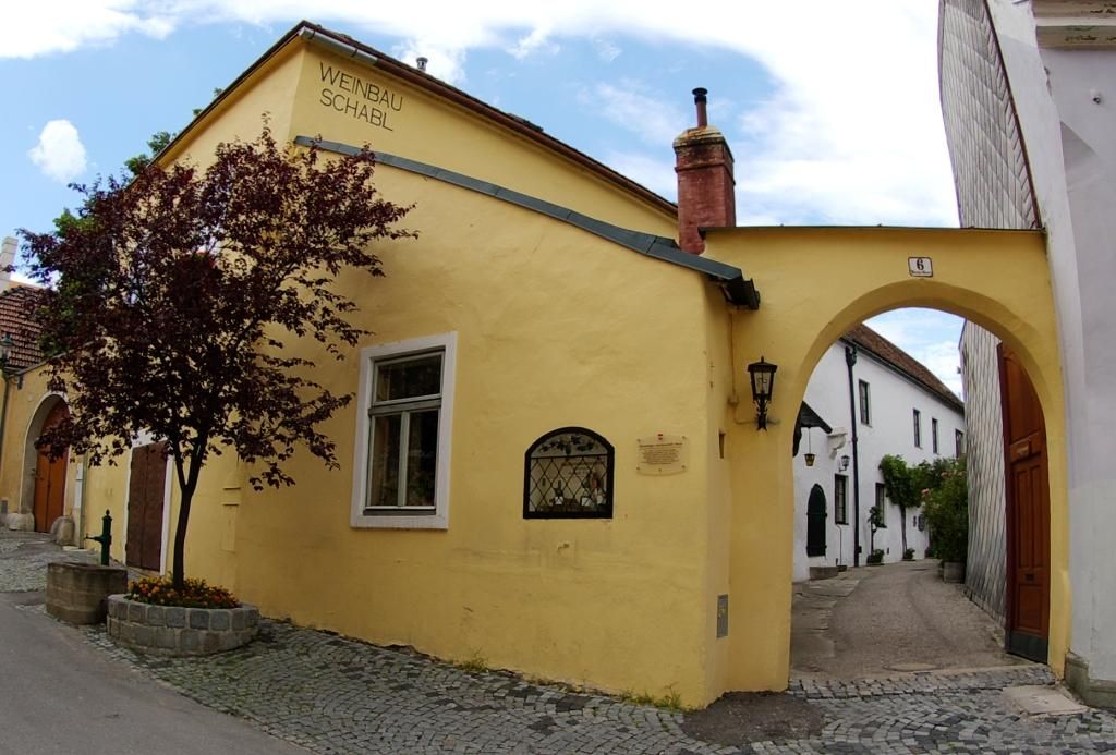 Žltá budova s nápisom "Weinbau Schabl", vedľa nej oblúk a strom.