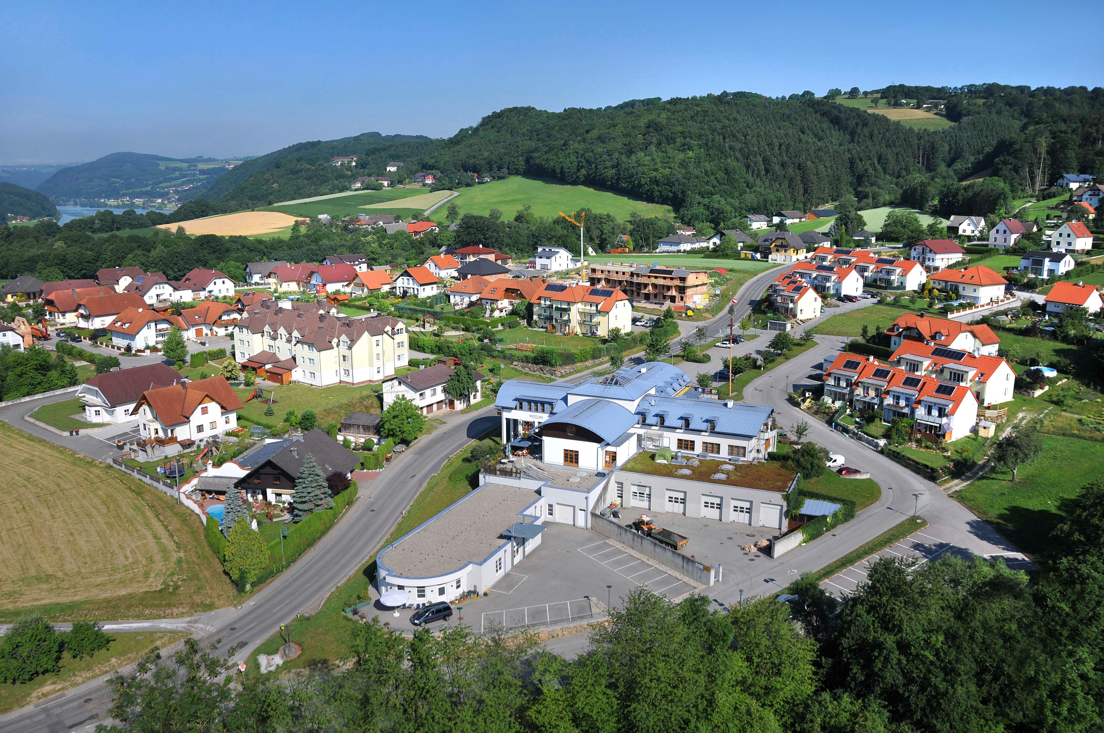 Letecký pohľad na obec s komunitným centrom a okolitými domami v zelenej kopcovitej krajine.