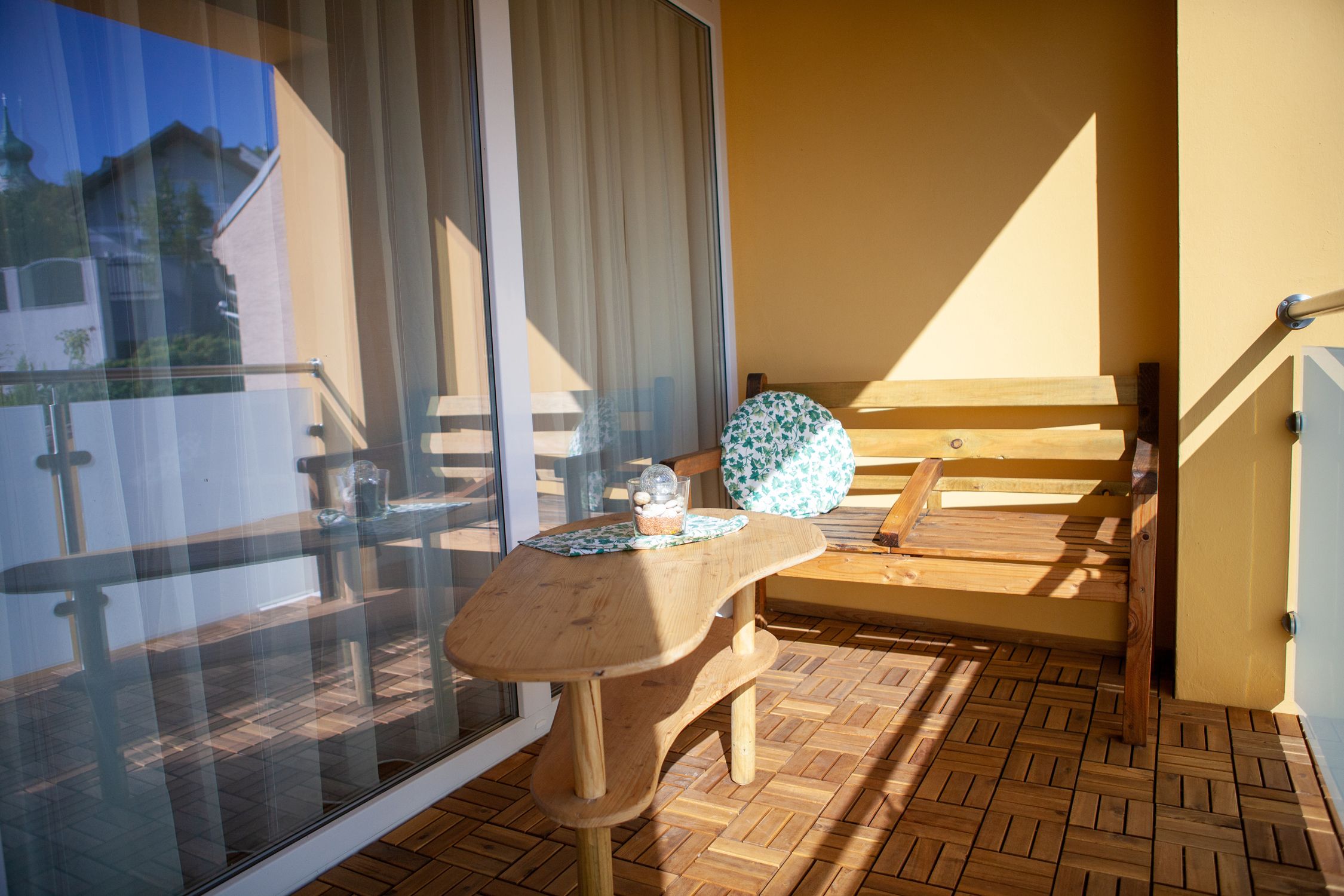 Balkón s drevenou podlahou, dreveným stolom a drevenou lavicou s okrúhlym vankúšom. V pozadí je vidieť sklenené zábradlie a budovu.