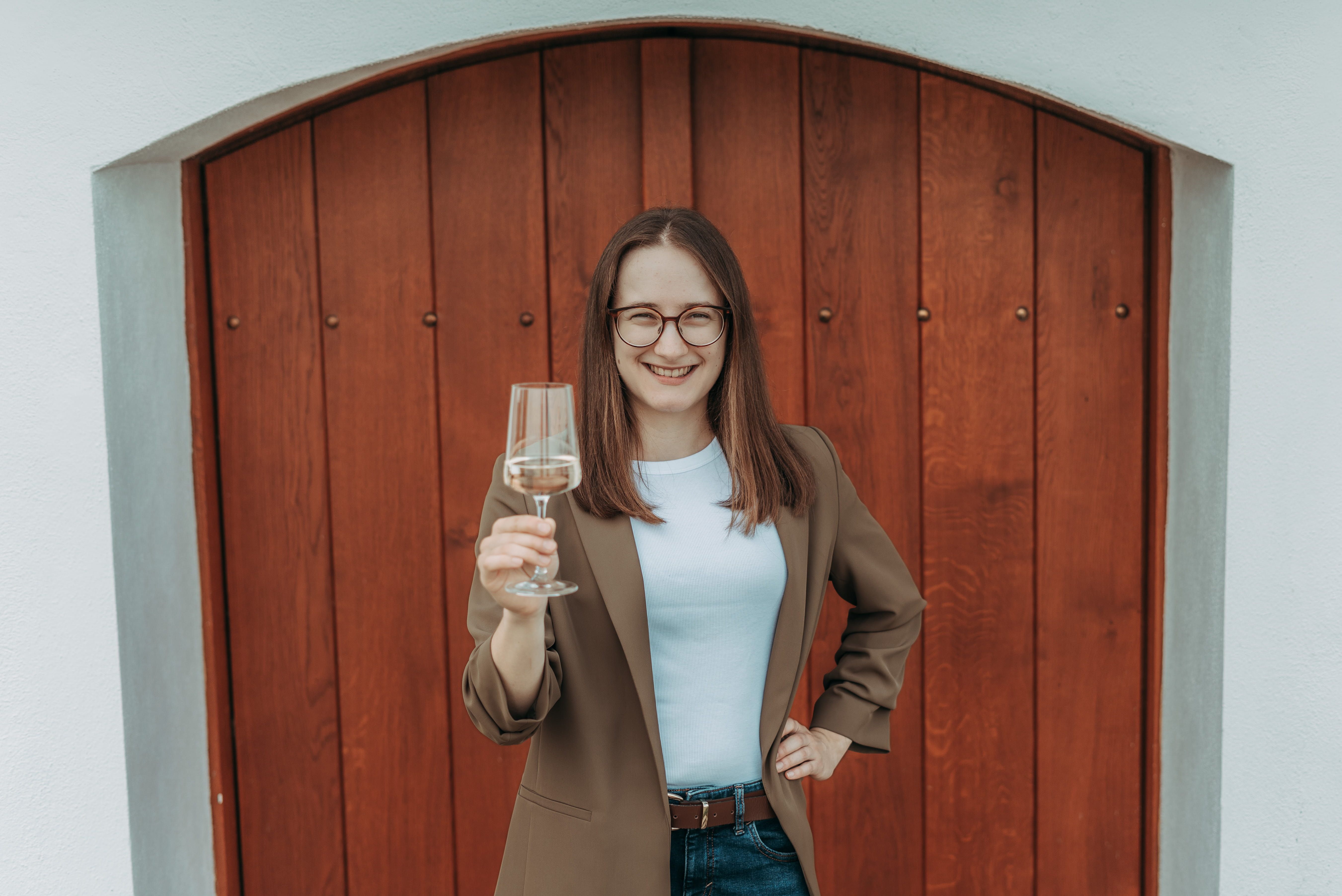 Žena s okuliarmi drží pohár na víno a usmieva sa pred drevenými dverami.