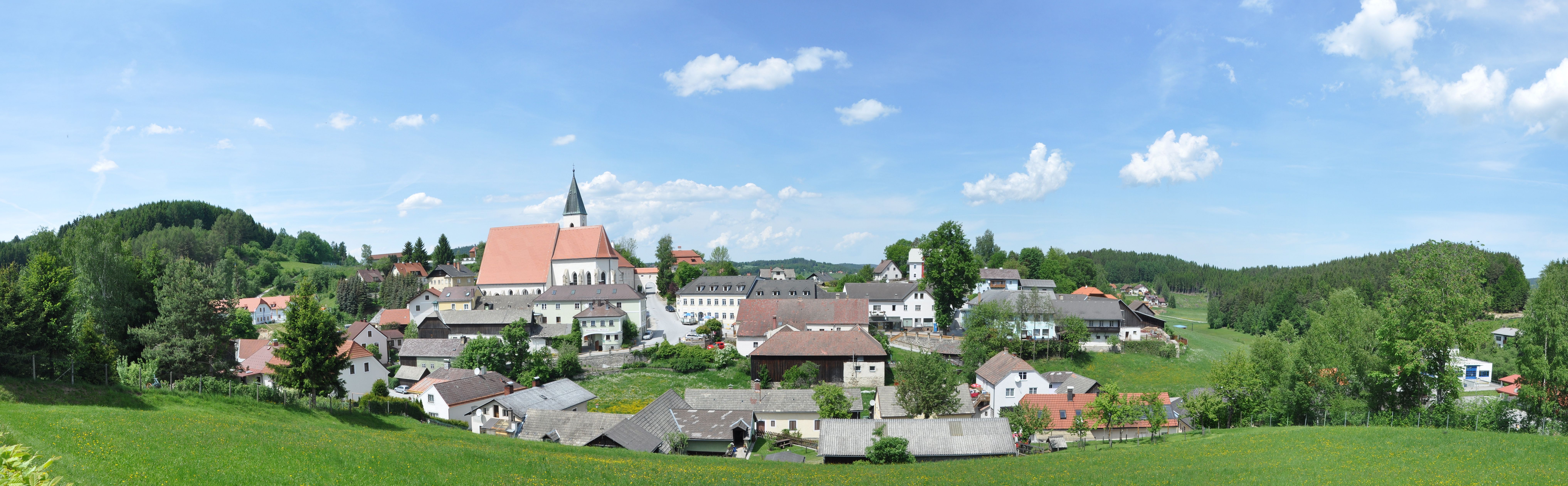 Panoráma trhového mestečka Schönbach s kostolom a okolitými domami, obklopeného zelenými lúkami a lesmi pod modrou oblohou.