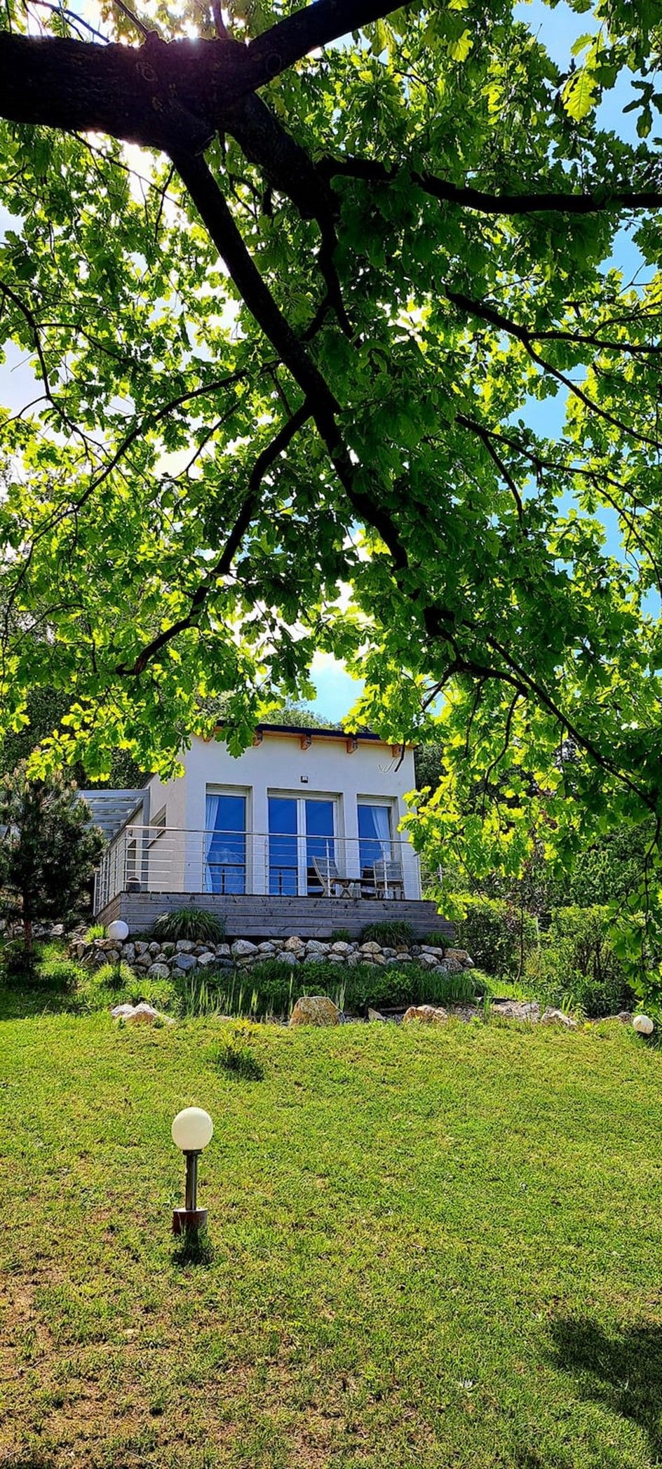 Moderná chata s veľkým predným oknom, obklopená zelenou prírodou a stromami.