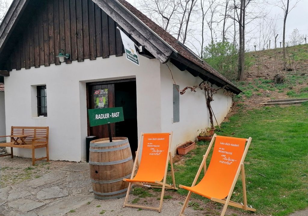 Malá biela budova s drevenou strechou, dvoma oranžovými lehátkami a dreveným sudom pred ňou, obklopená stromami a trávou.