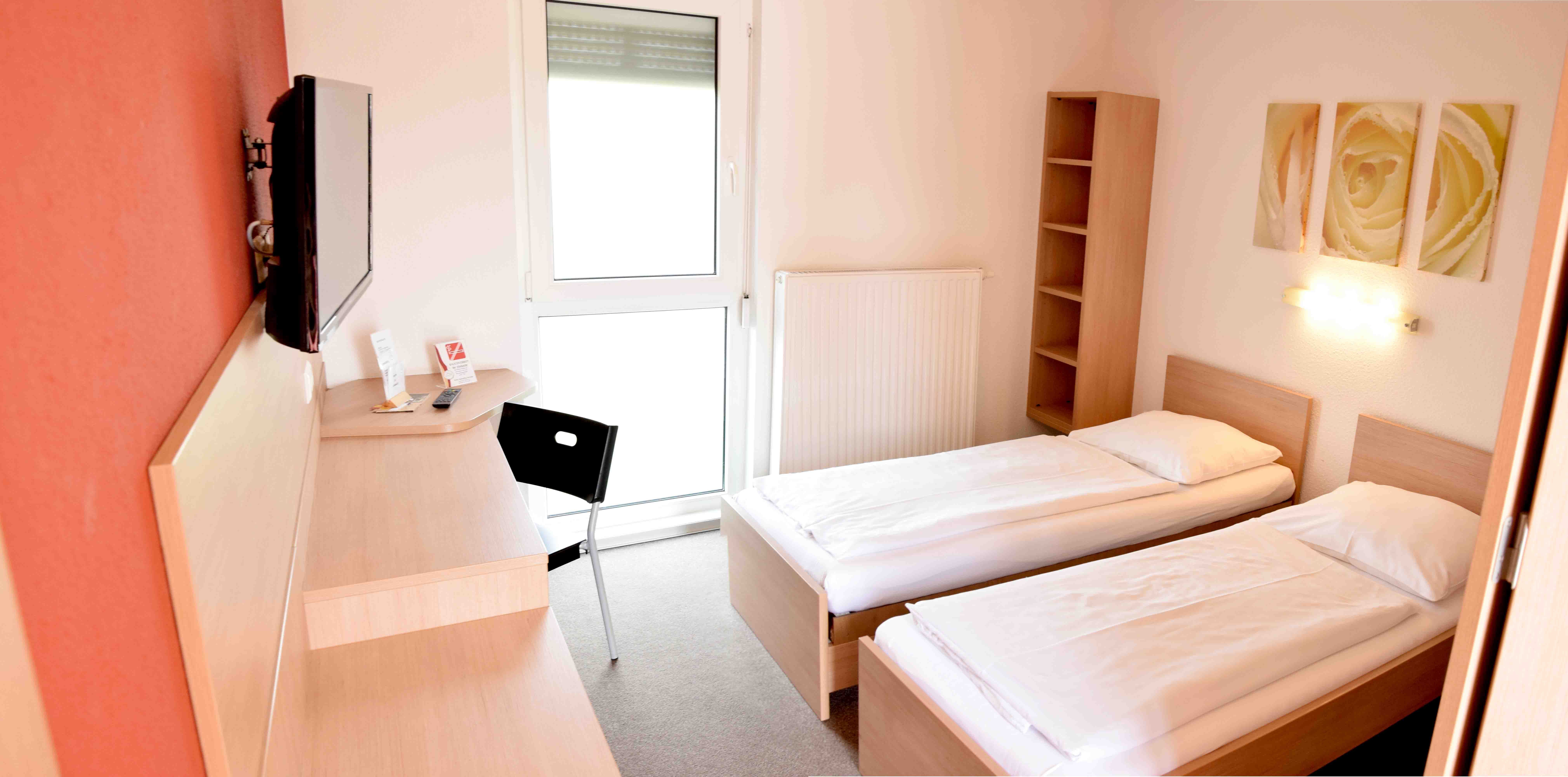 Jednoduchá motelová izba s dvoma samostatnými posteľami, stolom, stoličkou a televízorom na stene.