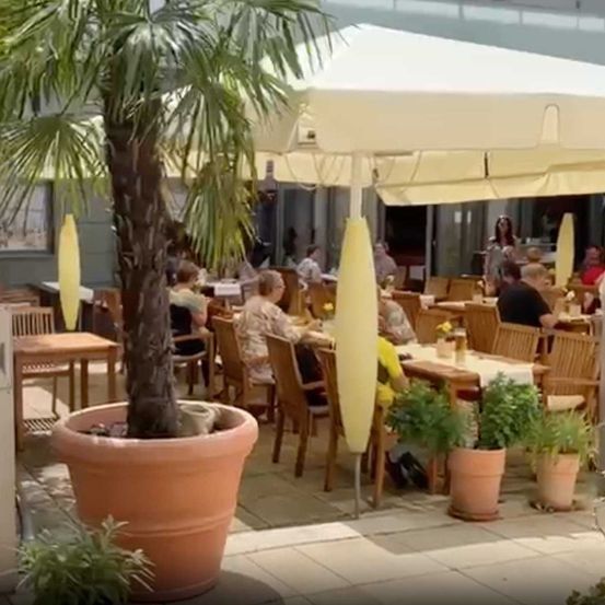 Vonkajšia časť kaviarne s drevenými stolmi a stoličkami, obklopená rastlinami a veľkým slnečníkom.