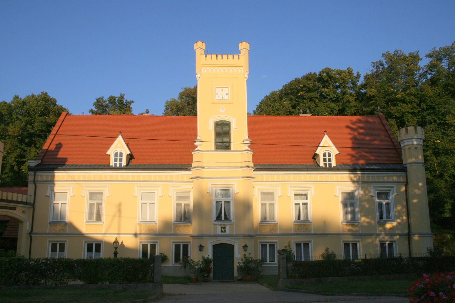 Žltý hrad s červenou strechou a vežou, obklopený stromami.
