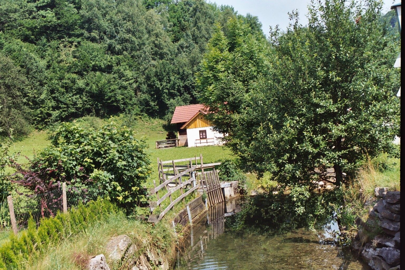 Malá chatka s červenou strechou stojí v zelenej krajine vedľa potoka a stromov.