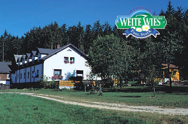 Biely statok s kvetmi pred lesom, nad ktorým je logo "Biohof Weite Wies Fam. Stark".