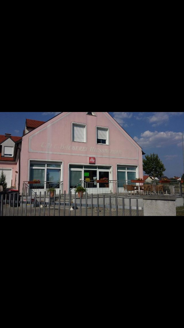 Pohľad zvonka na ružovú budovu s nápisom "Bäckerei-Café Hiessberger". Pred budovou sú stoly a stoličky.