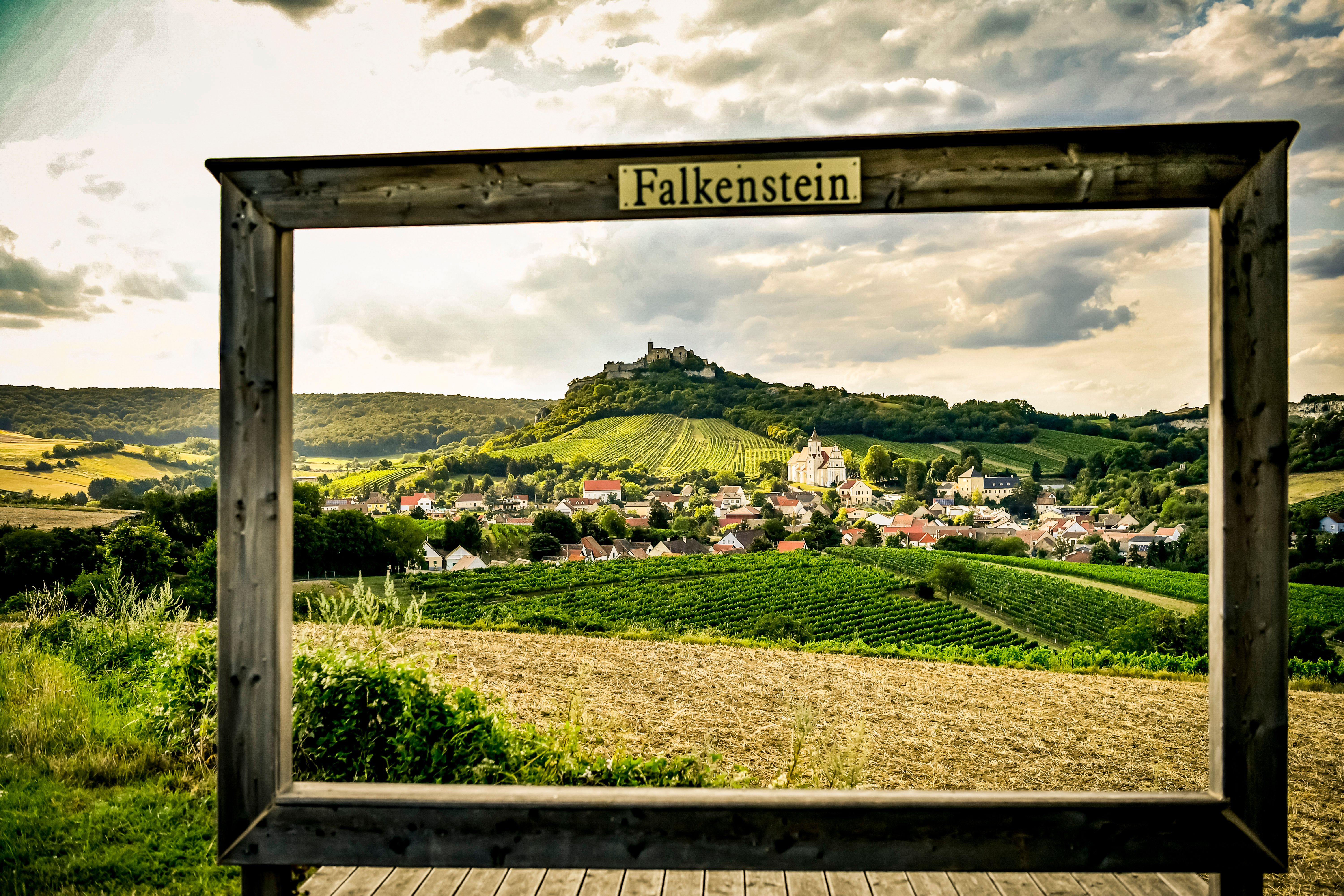 Pohľad na obec Falkenstein s vinicami a zrúcaninou hradu v pozadí, orámovaný dreveným rámom s nápisom "Falkenstein".