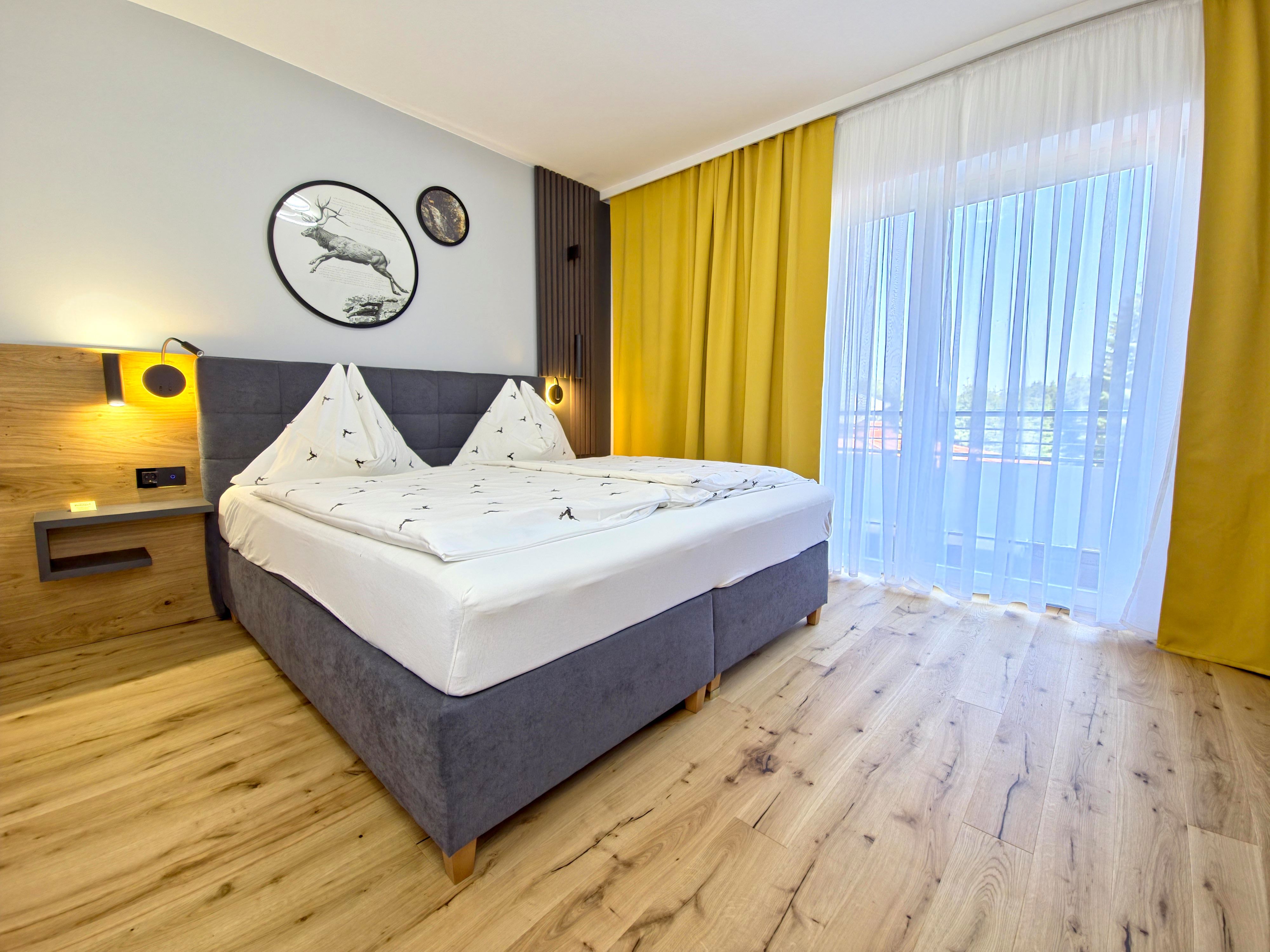 Moderná spálňa s manželskou posteľou, drevenou podlahou a žltými závesmi.