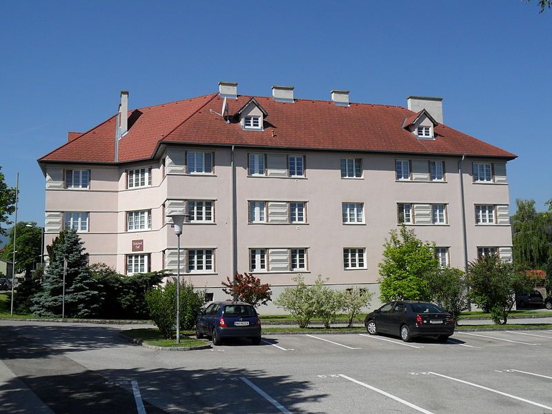 Obytná budova s červenou strechou a niekoľkými oknami, obklopená stromami a zaparkovanými autami.