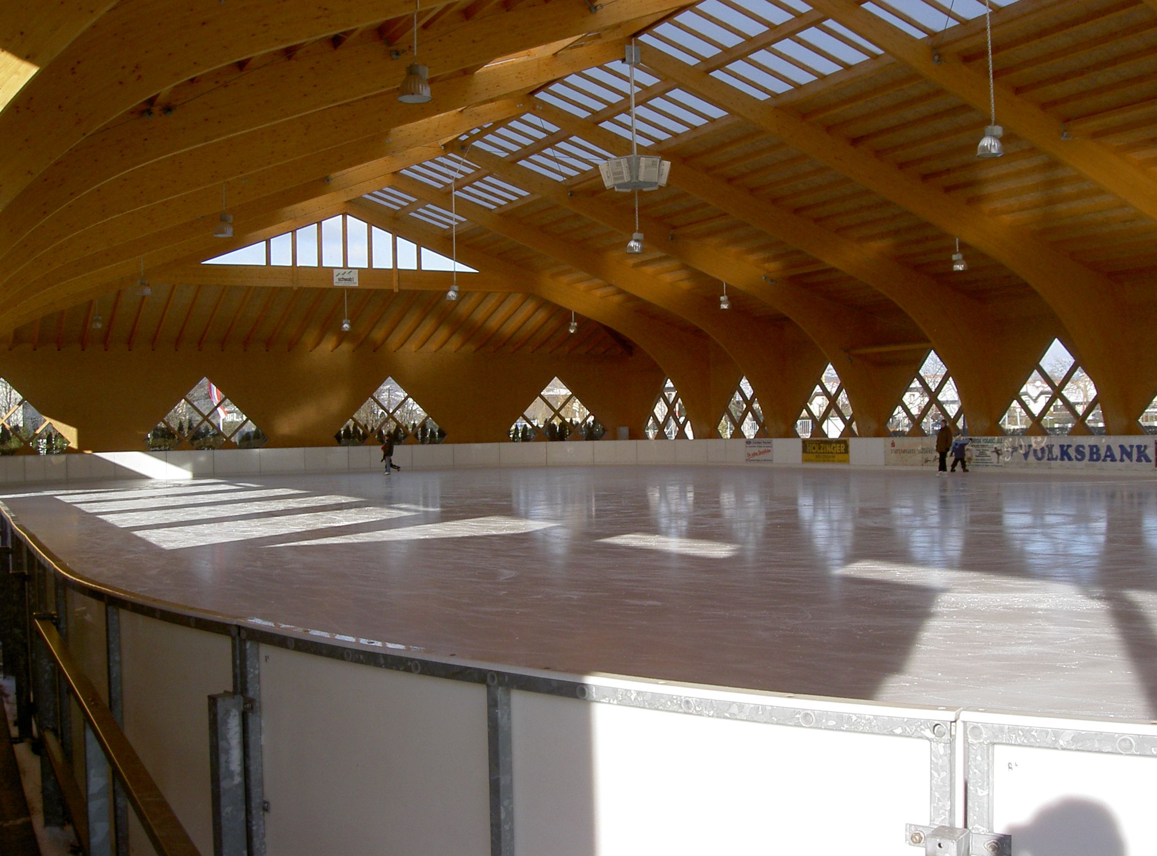 Pohľad do interiéru krytej umelej ľadovej plochy s drevenou strechou a niekoľkými ľuďmi na ľade.