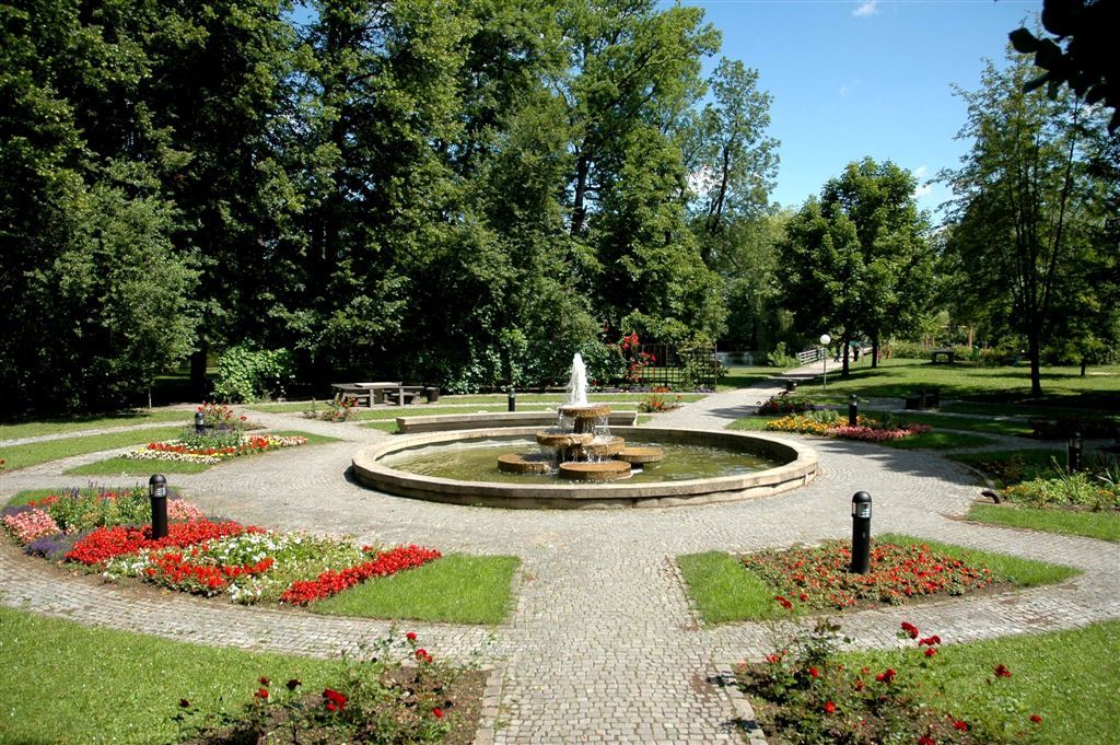 Upravený park s centrálnou fontánou, obklopený kvetinovými záhonmi a stromami.