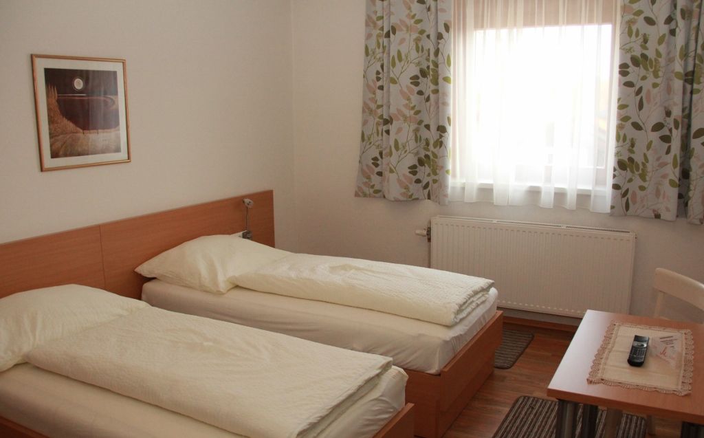Izba pre hostí s dvoma samostatnými posteľami, oknom so závesmi, stolíkom s diaľkovým ovládaním a obrazom na stene.