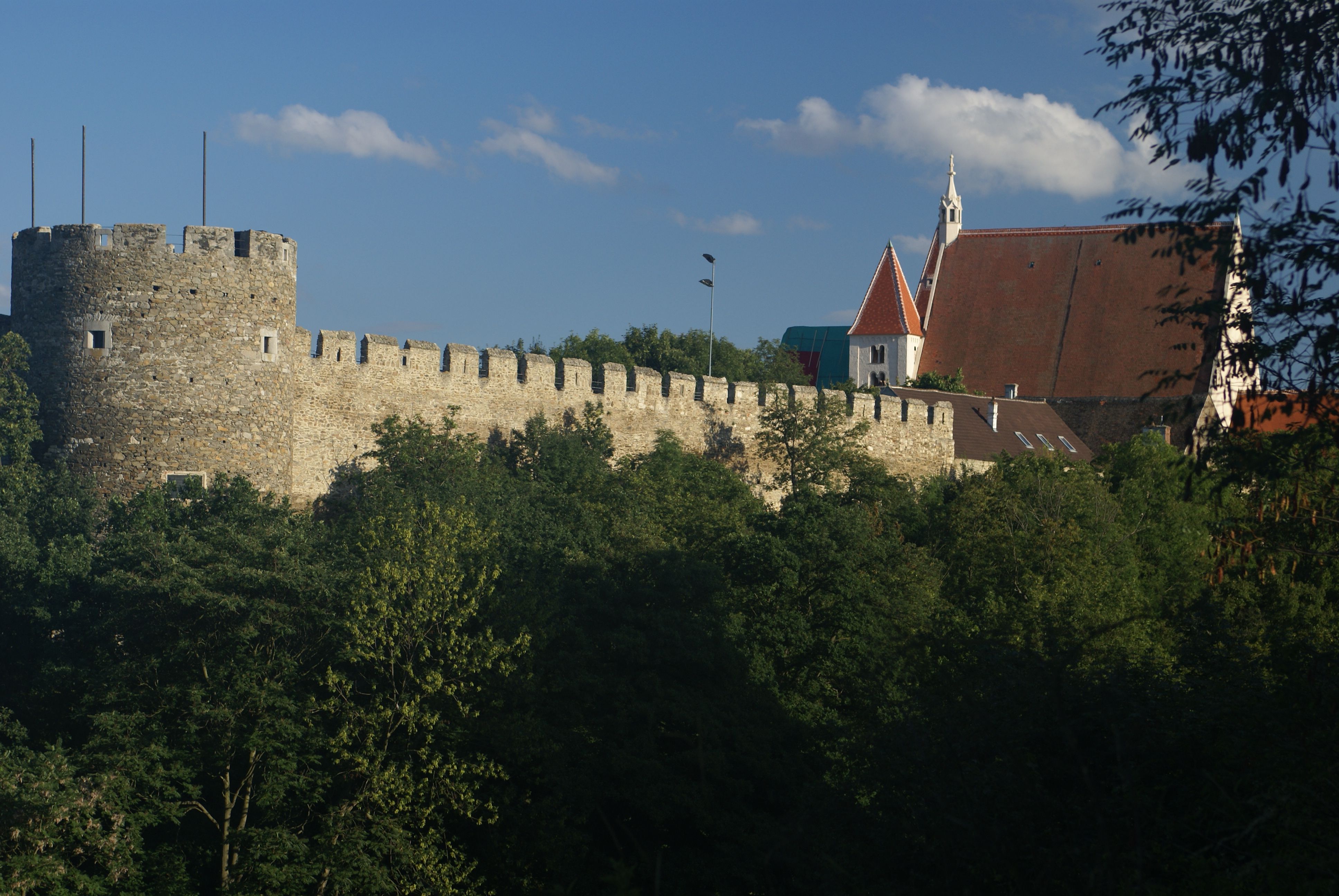 Mestské hradby s kancelárskou vežou a farským kostolom v pozadí, obklopené stromami.