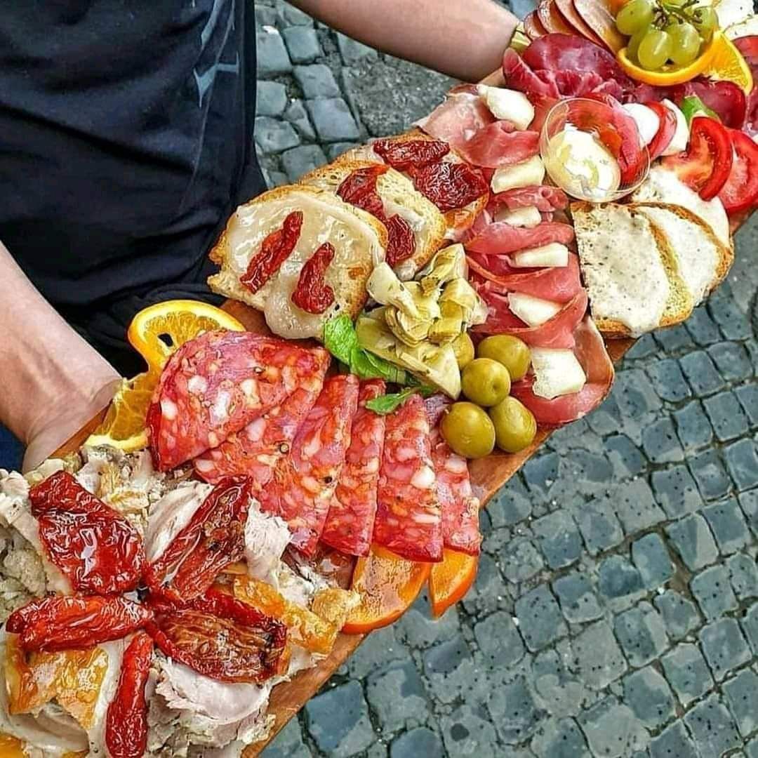 Jedna osoba drží v ruke veľký drevený tanier s rôznymi antipasti vrátane klobásy, syra, olív a sušených paradajok.
