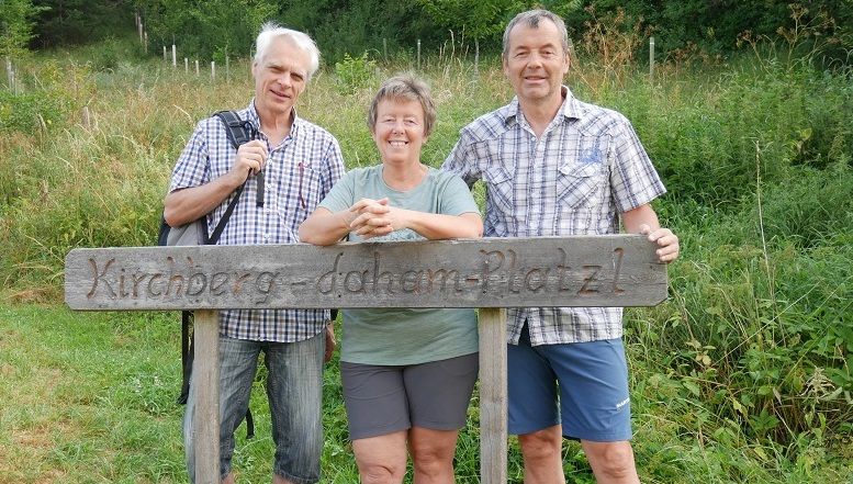 Tri osoby stoja za drevenou tabuľou s nápisom "Kirchberg-daham-Platzl".