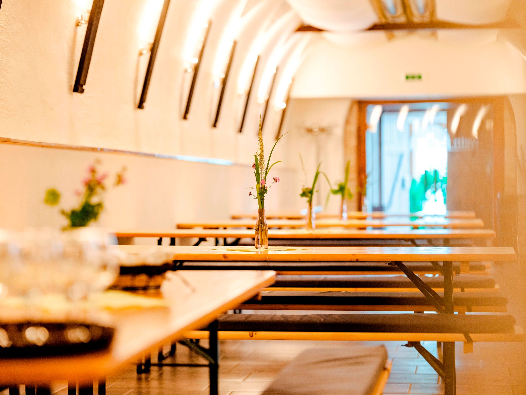 Pohľad do interiéru vínneho šenku s dlhými drevenými stolmi a lavicami, zdobenými vázami s kvetmi.