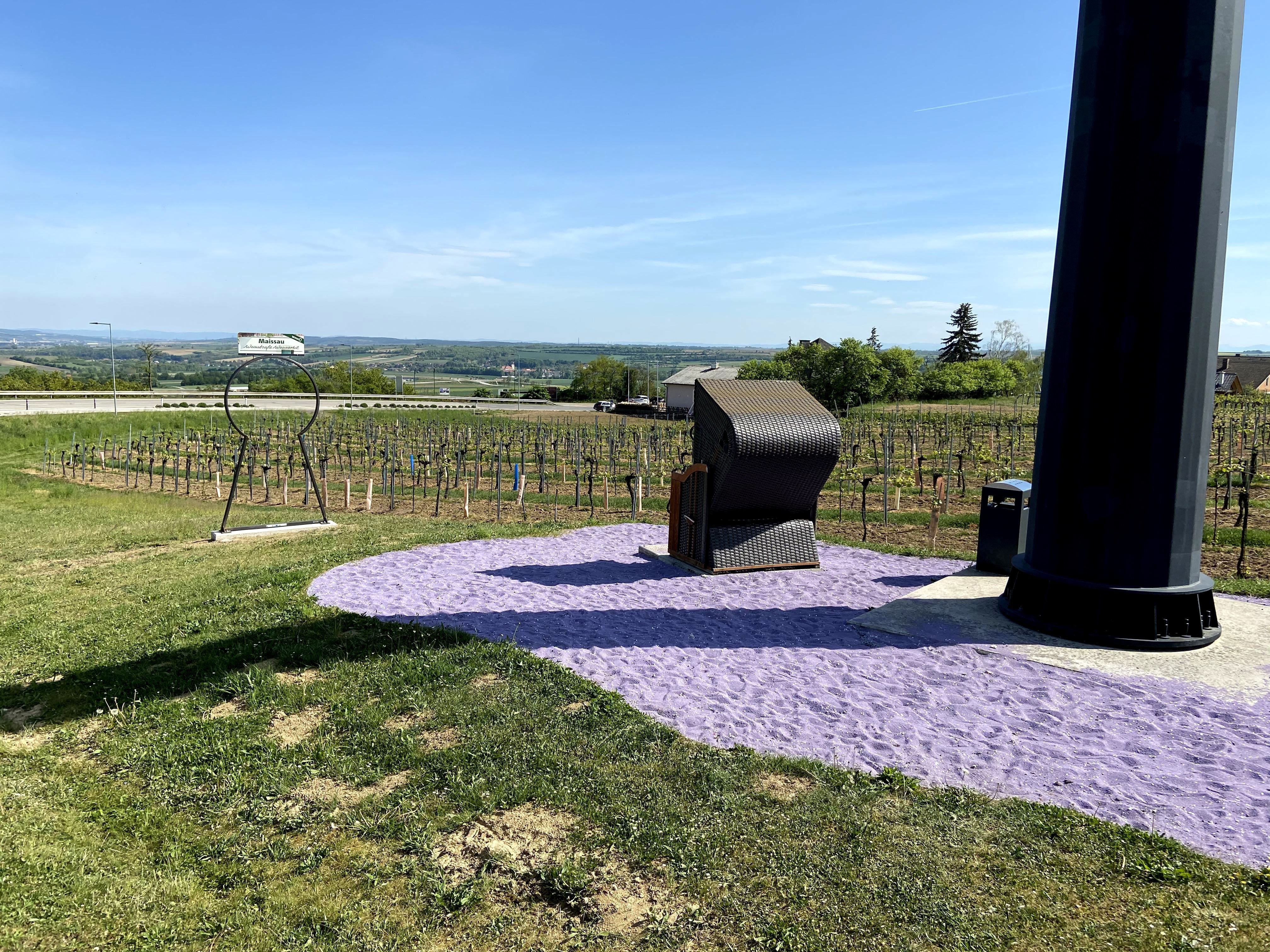 Plážové ležadlo stojí na fialovom piesku vedľa veľkého čierneho stĺpa, obklopené vinicami a rozľahlou krajinou.