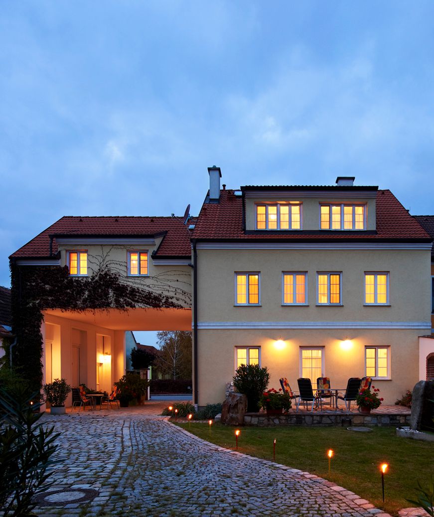 Osvetlená budova s vydláždenou cestou a záhradným nábytkom v popredí.