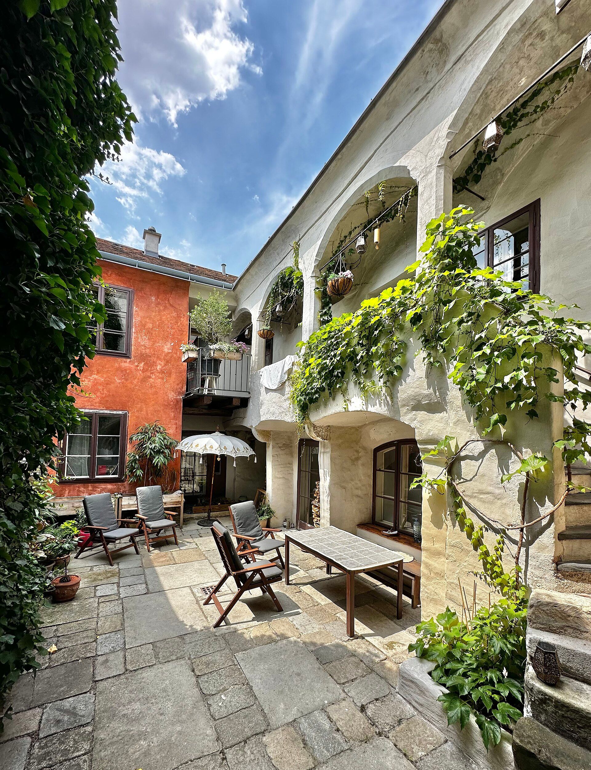 Útulný dvor s rastlinami, dreveným nábytkom a slnečníkom.