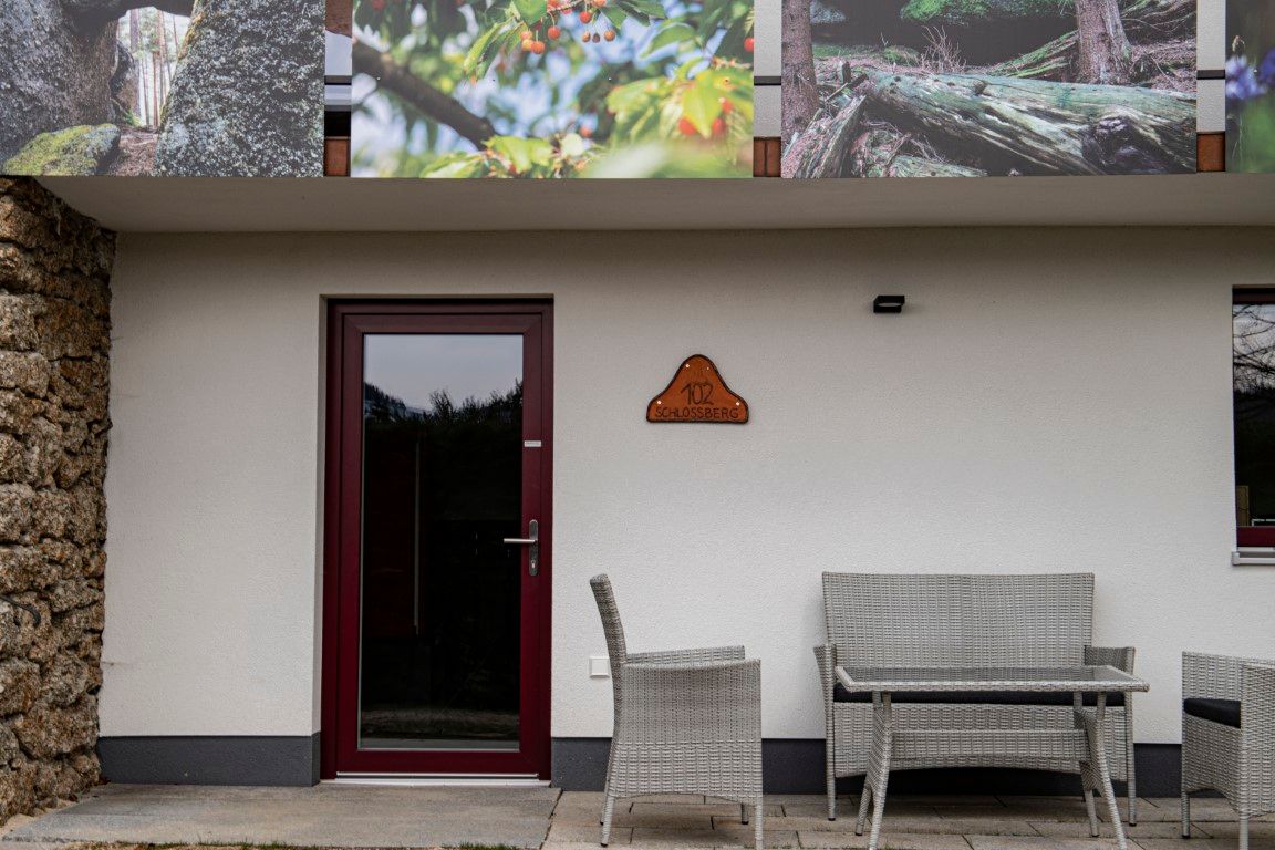Vstupná časť budovy s dverami, tabuľkou s nápisom „102 Schlossberg“ a záhradným nábytkom pred ňou.