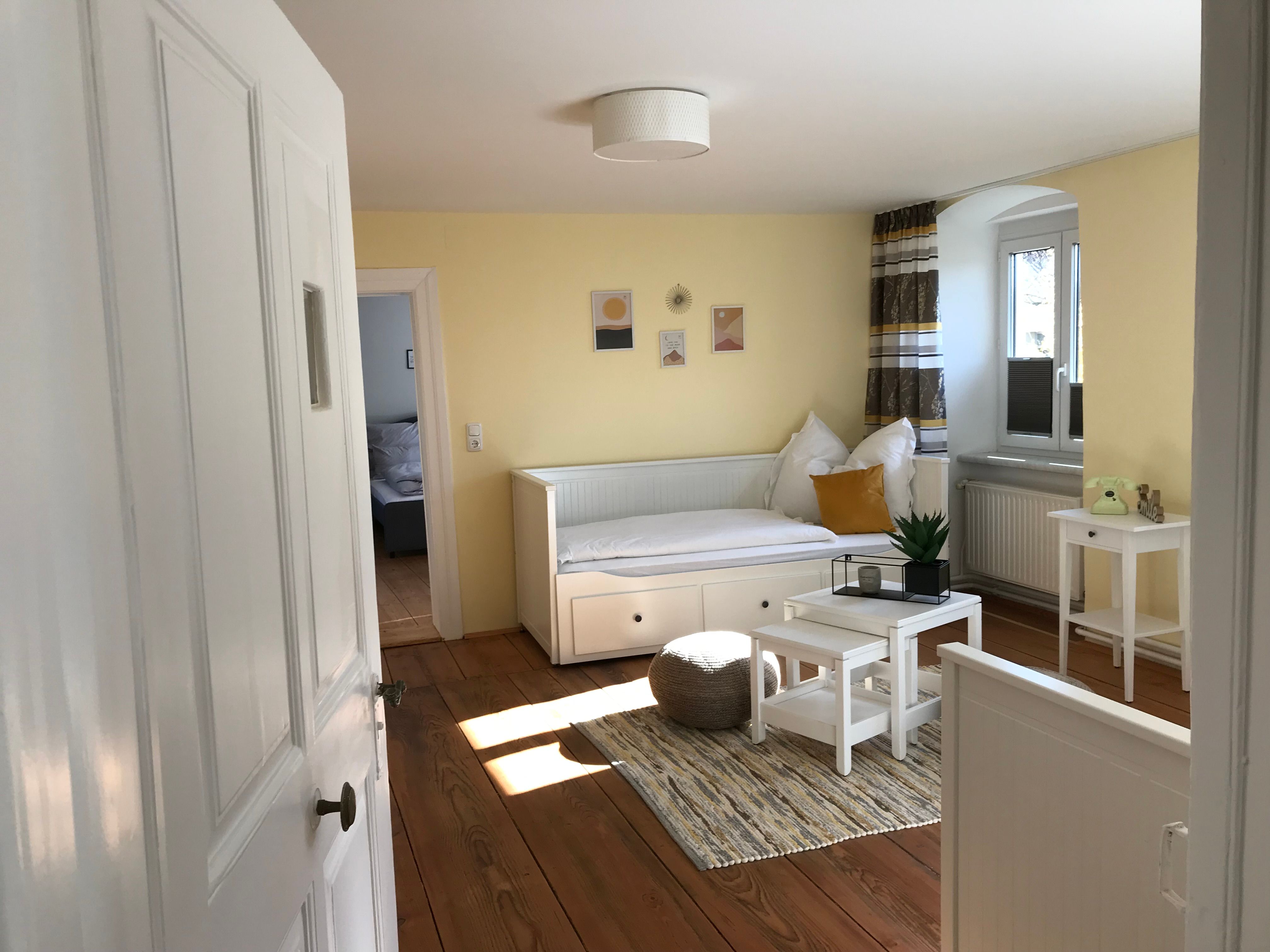 Svetlá izba so žltými stenami, bielou dennou posteľou, stolíkom a rastlinami.