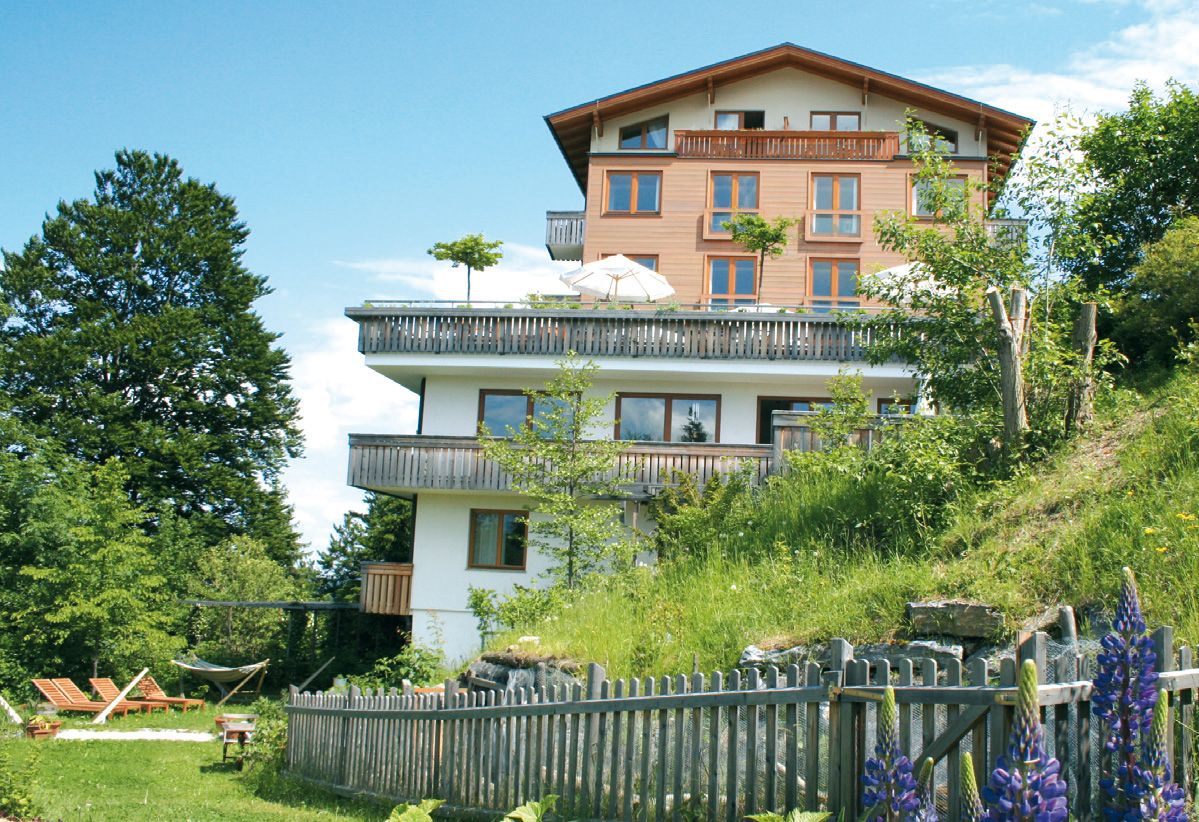 Viacposchodový hotel s drevenými balkónmi, obklopený zelenou prírodou a kvitnúcimi kvetmi, pod modrou oblohou.