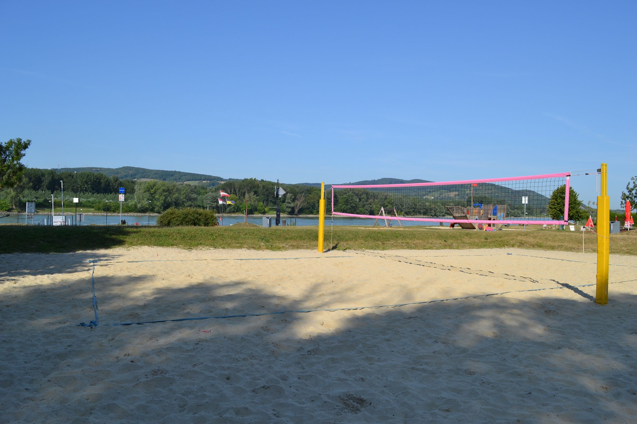 Prázdne ihrisko na plážový volejbal so žltými stĺpikmi a ružovou sieťou pred riekou a zelenými kopcami v pozadí.