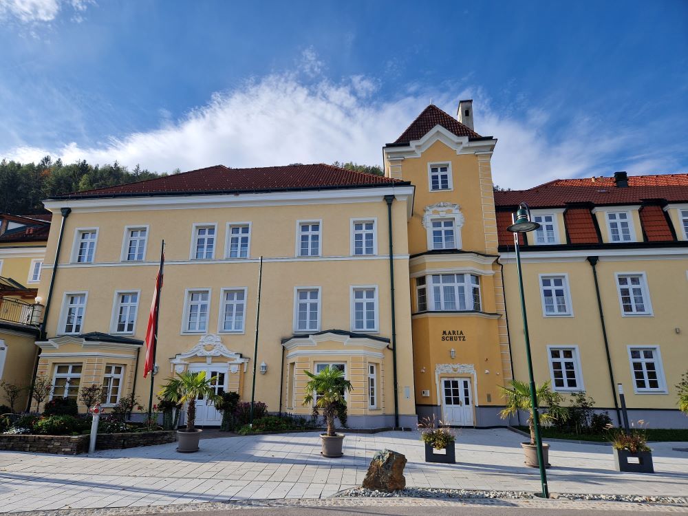 Žltá budova s červenými strechami a niekoľkými oknami, obklopená rastlinami a vlajkami.