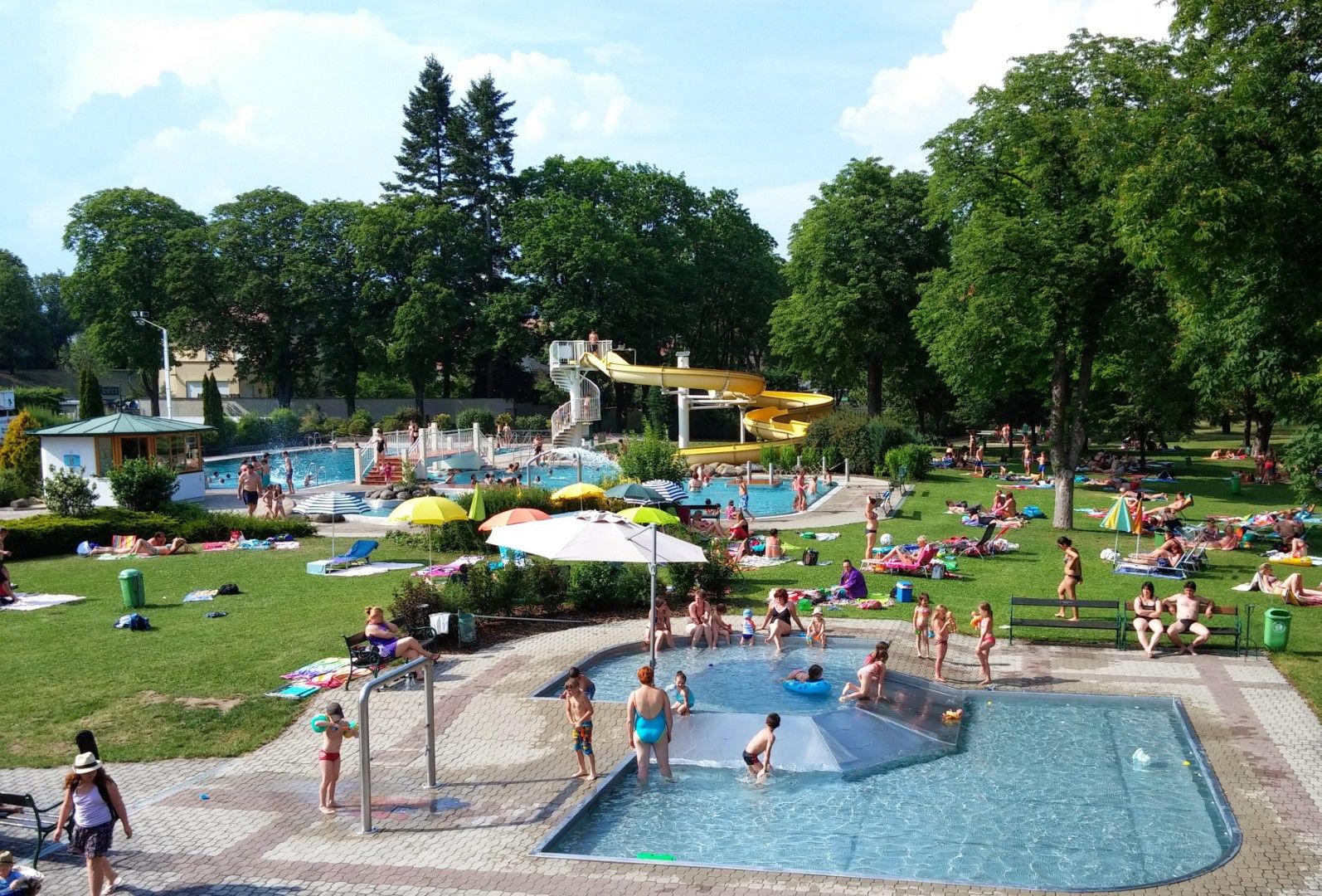 Vonkajší bazén s niekoľkými bazénmi, žltou šmykľavkou a množstvom ľudí relaxujúcich na trávniku.