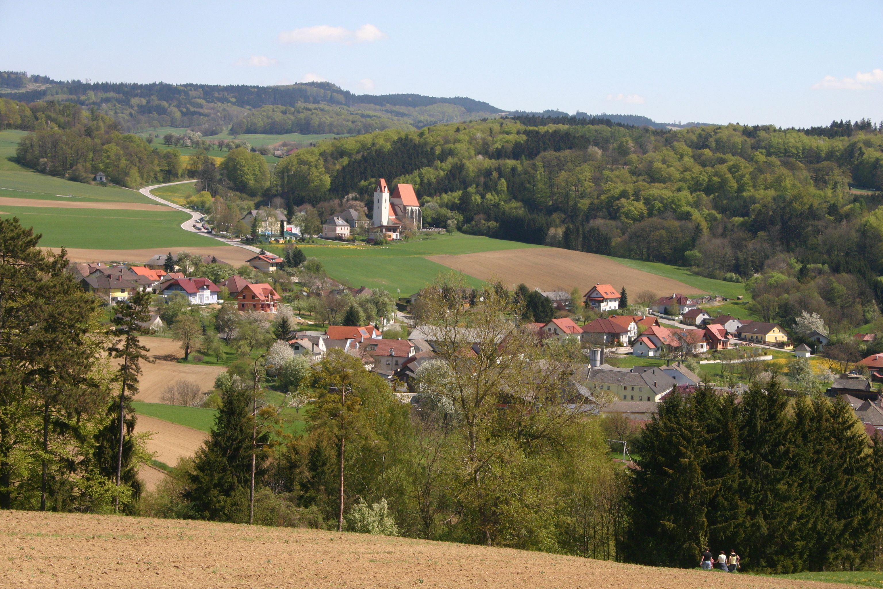 Krajina s dedinou, kostolom a kopcami v pozadí.