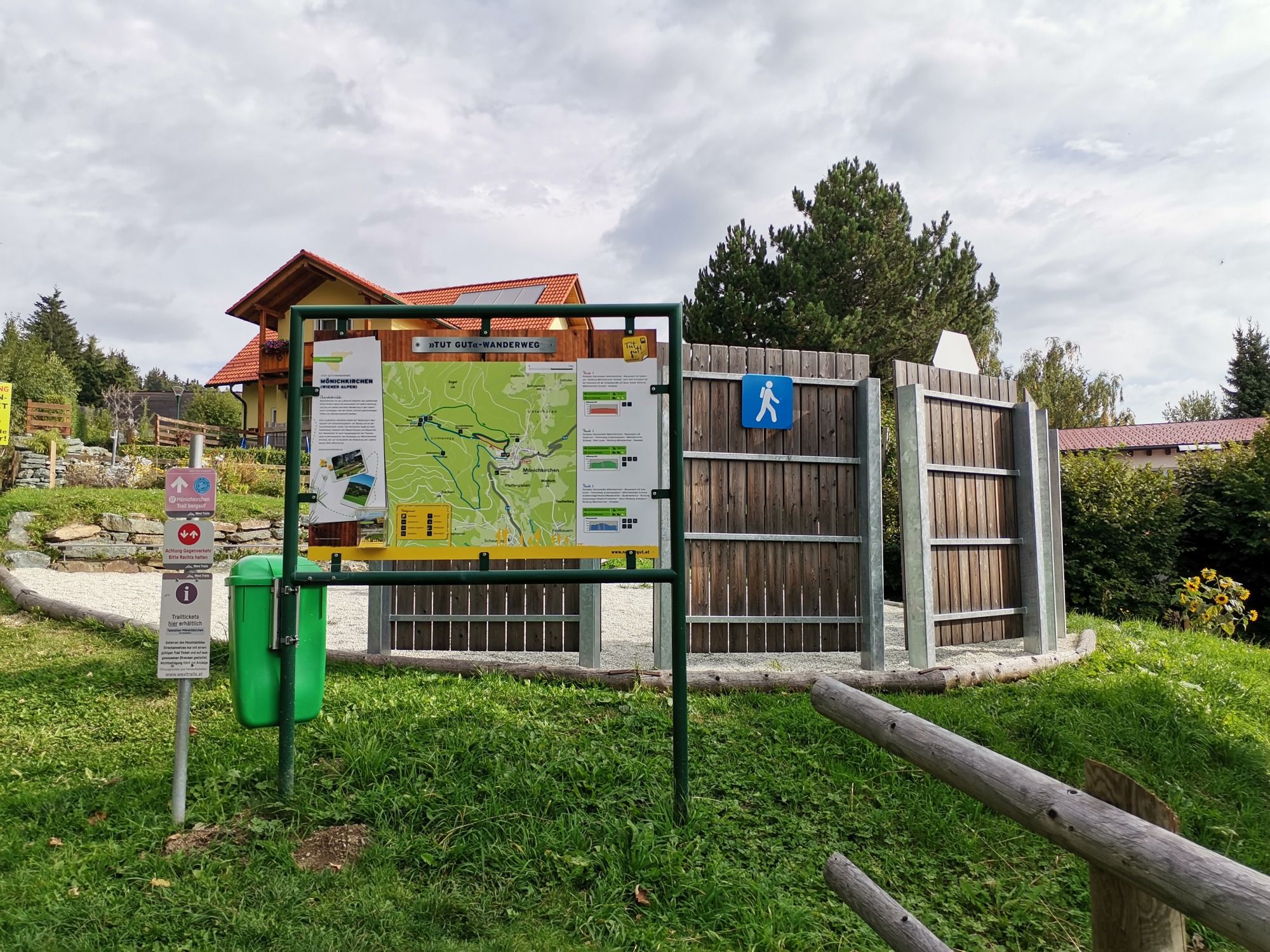 Informačná tabuľa turistického chodníka s mapou a značkami na lúke, obklopená stromami a budovou v pozadí.