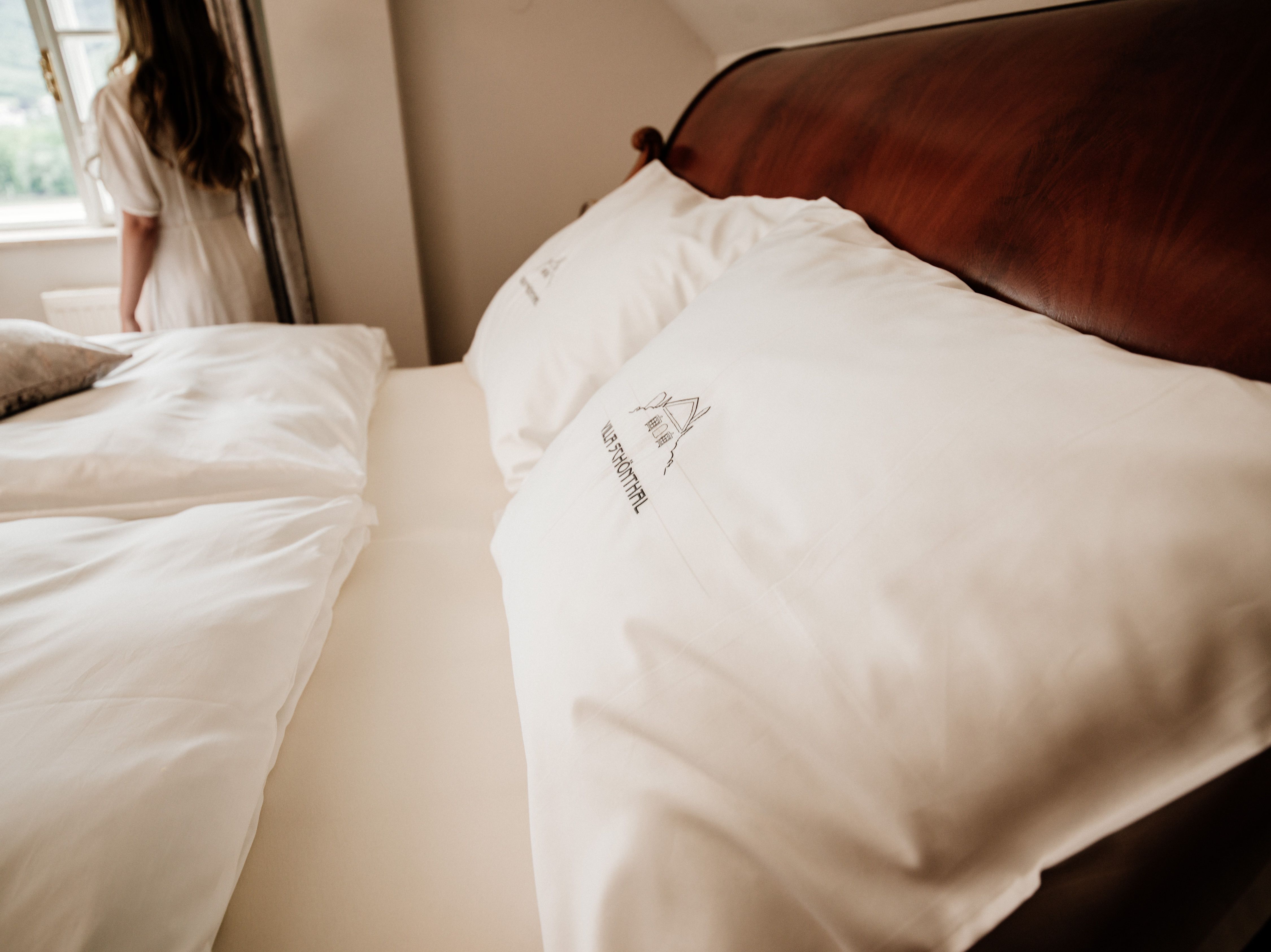 Detailný záber na posteľ s bielymi vankúšmi a posteľnou bielizňou, v pozadí žena pri okne.