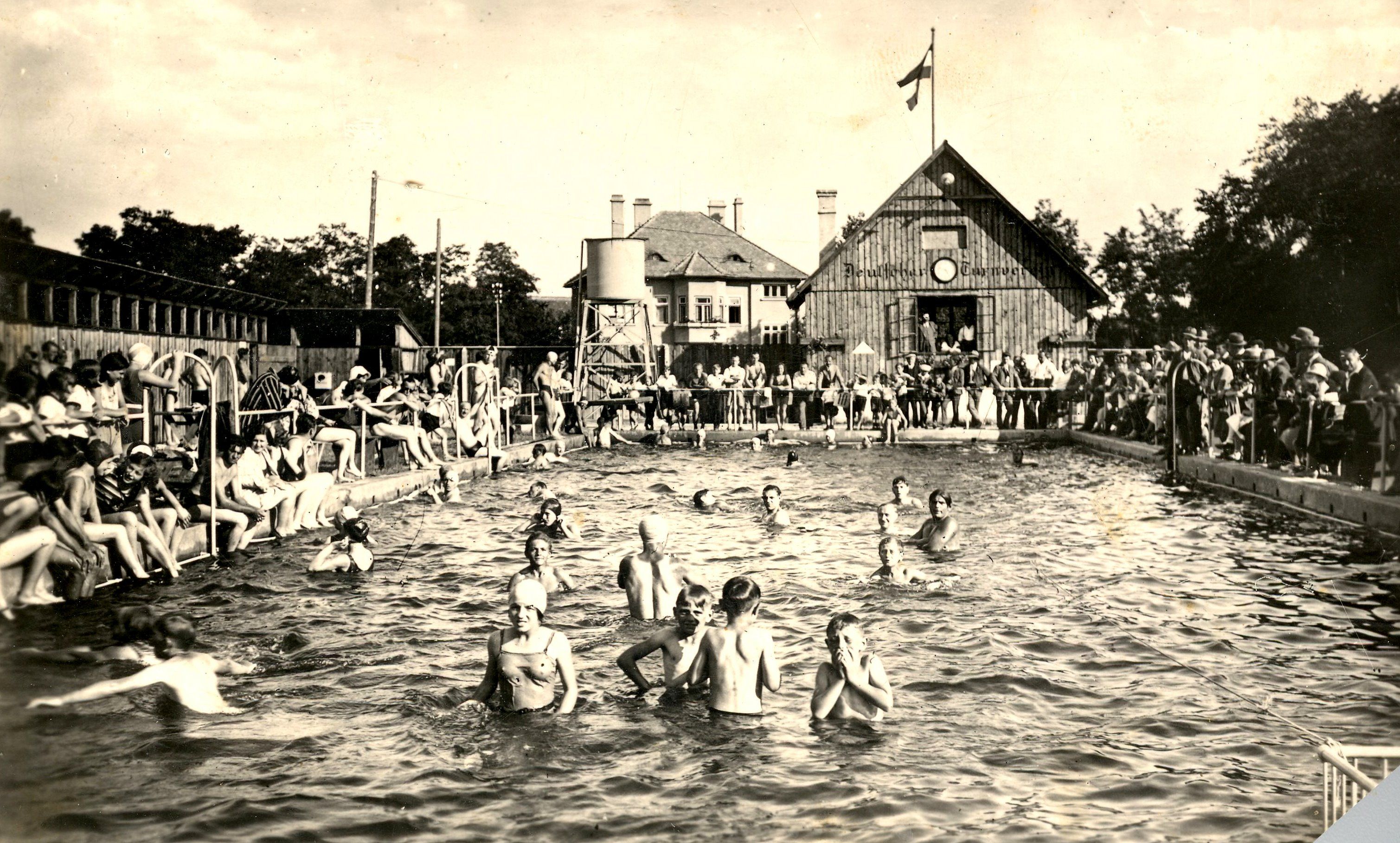 Historická fotografia vonkajšieho kúpaliska s množstvom ľudí vo vode a na okraji bazéna. V pozadí je budova s vlajkou.