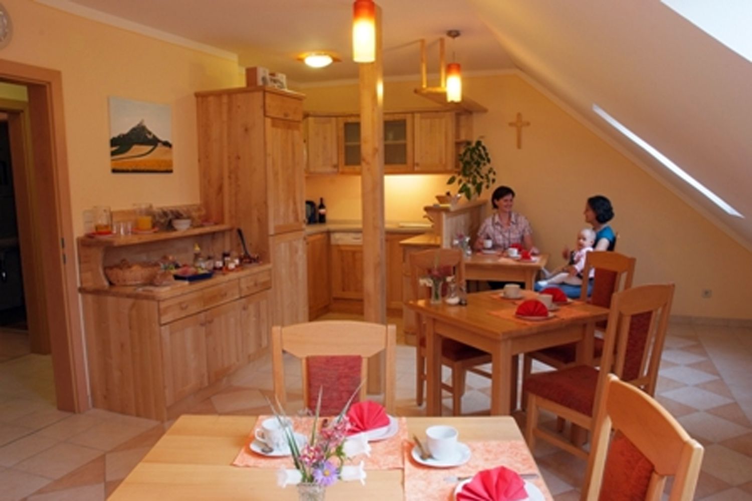 Útulná raňajková miestnosť s dreveným nábytkom, prestretými stolmi a dvoma rozprávajúcimi sa ľuďmi. Jednej osobe sedí na kolenách dieťa.