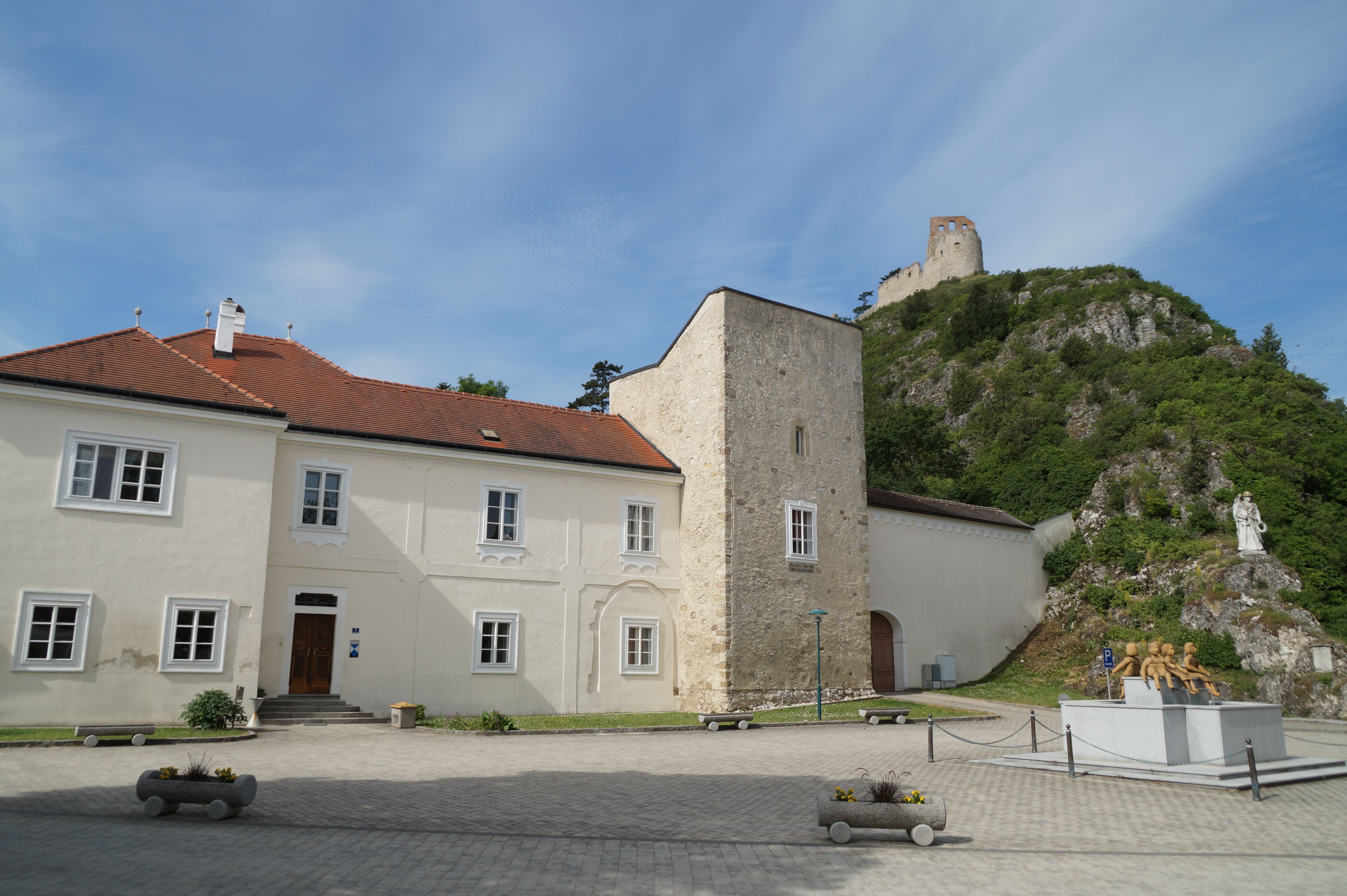 Historická budova s červenou škridlovou strechou a kamennou vežou, so zrúcaninou hradu na kopci v pozadí.