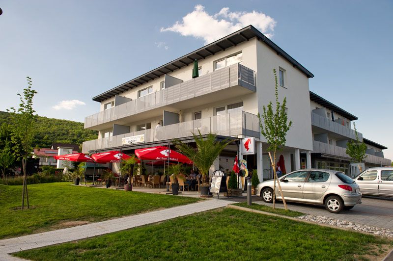 Pohľad zvonku na modernú budovu s kaviarňou a červenými slnečníkmi.