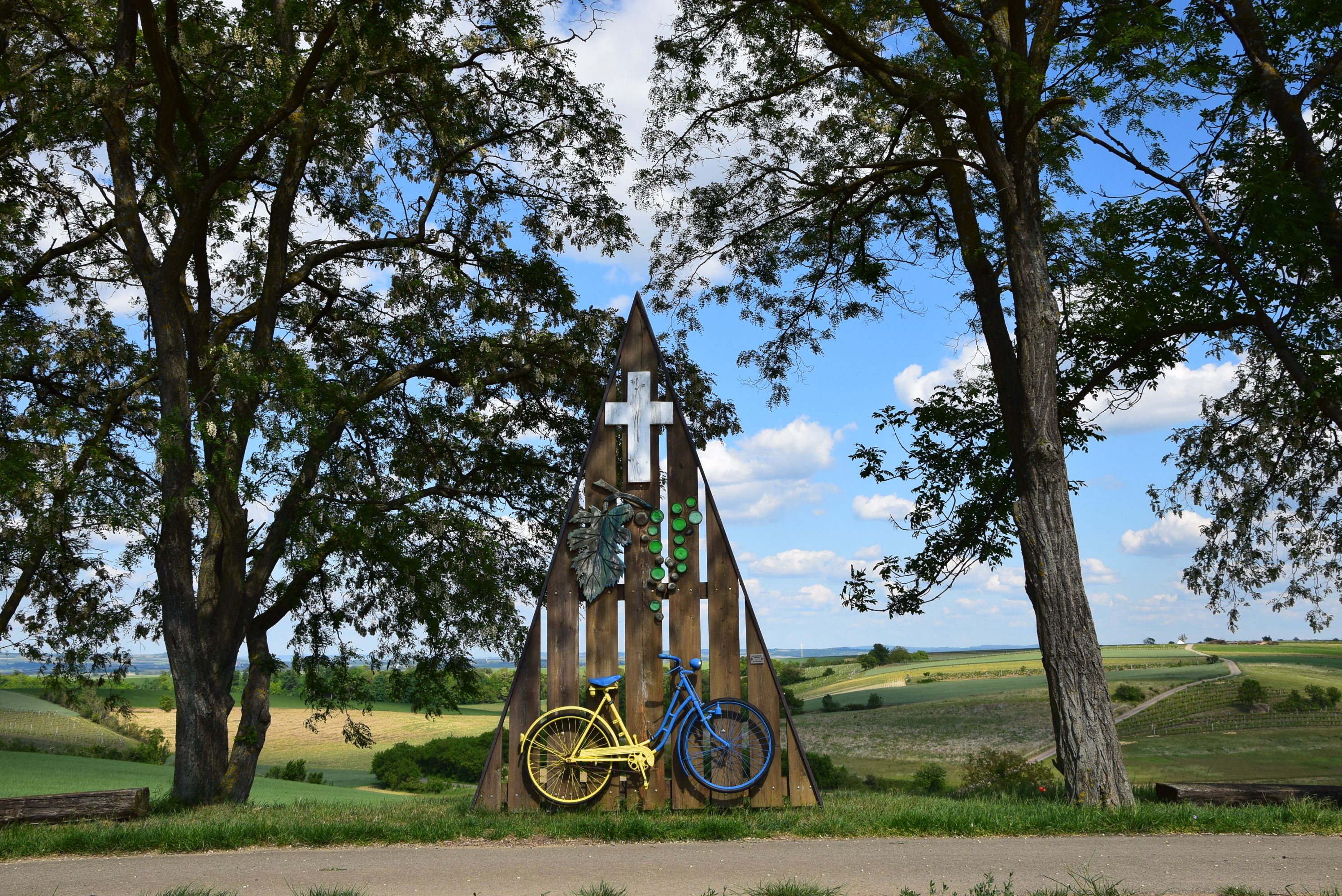 Svätyňa so žltým a modrým bicyklom, obklopená stromami a poľami pod modrou oblohou.