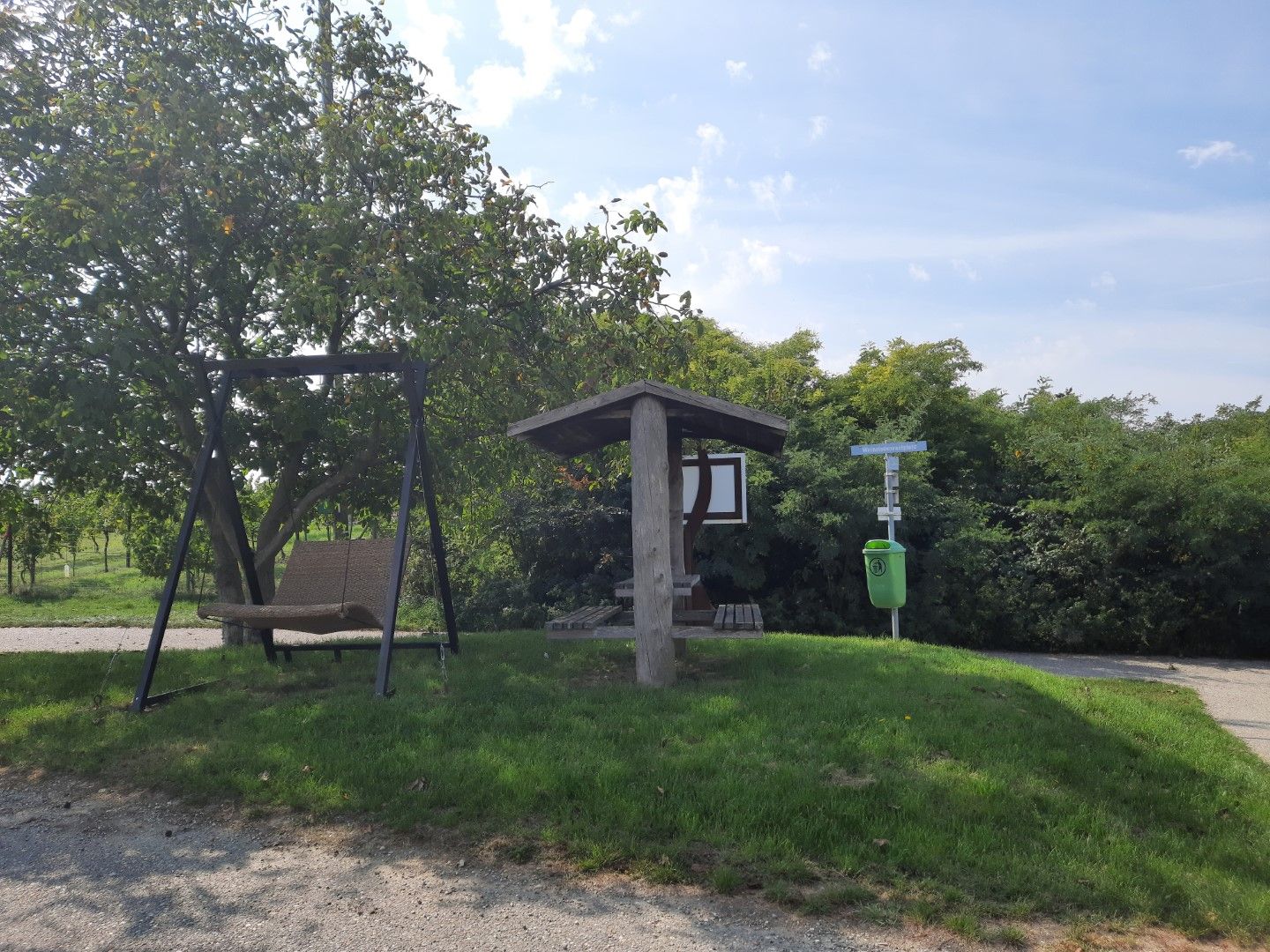Odpočívadlo s hojdačkou a krytým sedením pri strome a odpadkovom koši na lúke.