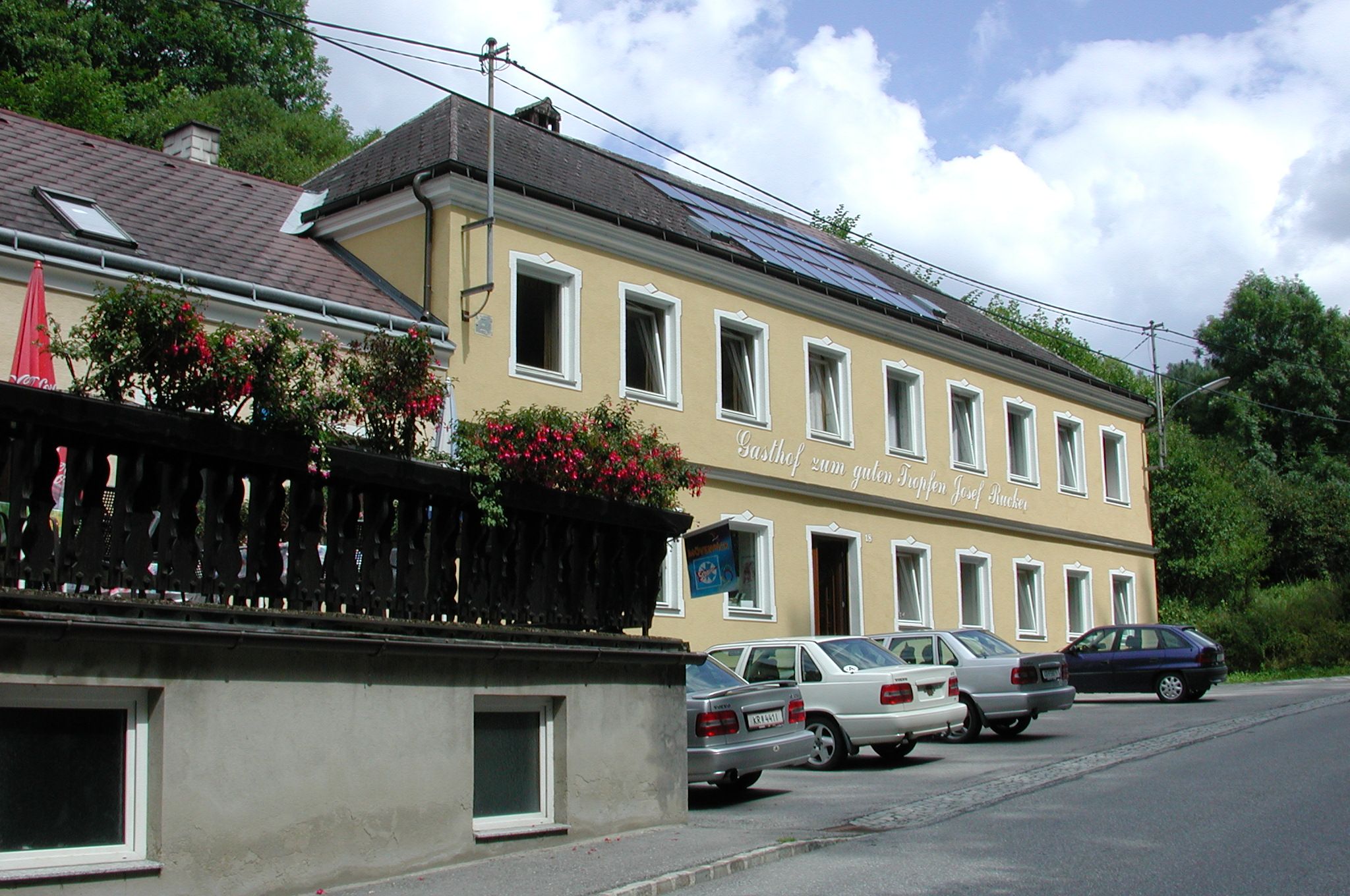 Žltá budova s nápisom "Gasthof zum guten Tropfen Josef Rucker", pred ňou zaparkované autá, v pozadí modrá obloha.
