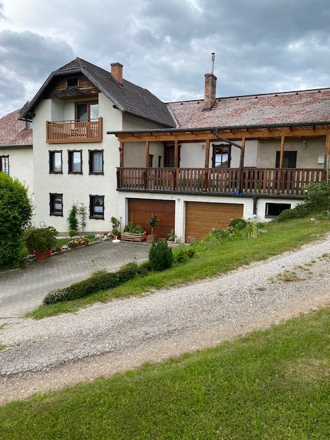 Dvojpodlažný dom s balkónom a garážou, obklopený zelenou krajinou a zamračenou oblohou.