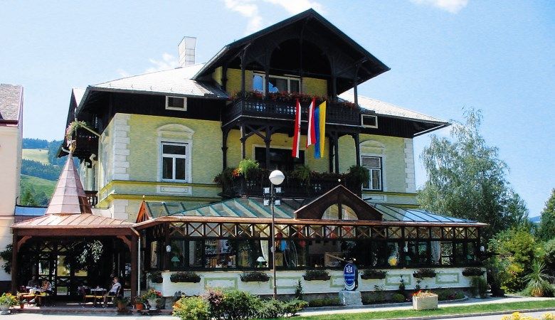 Tradičná budova s presklenou verandou a rakúskou vlajkou, obklopená rastlinami.