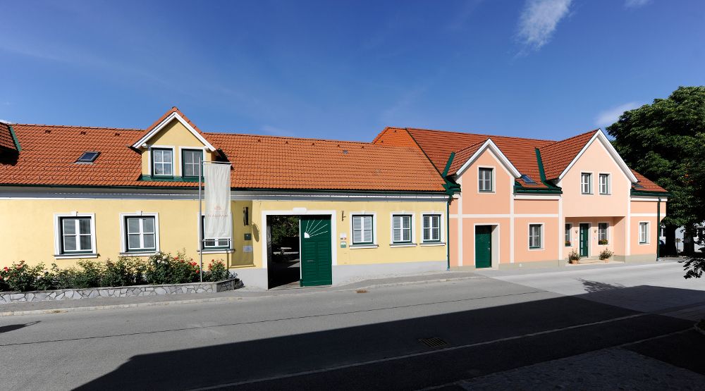 Dlhý jednoposchodový dom so žltou a ružovou fasádou, zelenými okenicami a červenou škridlovou strechou pod modrou oblohou.