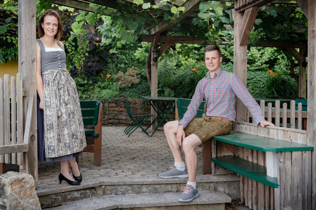 Muž v lederhosen a žena v dirndli pózujú v záhrade s drevenými konštrukciami.