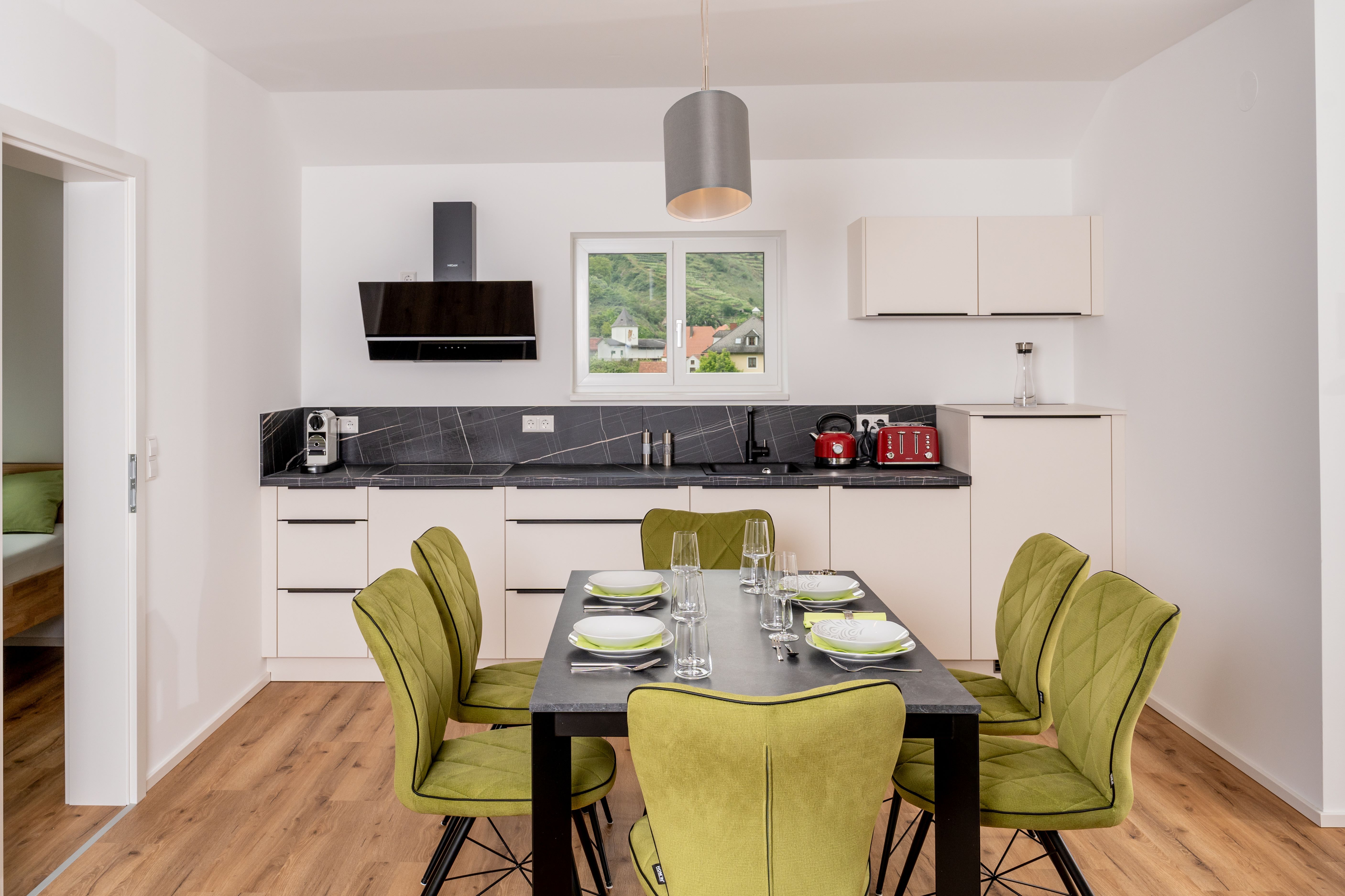 Moderná kuchyňa s jedálenským stolom a zelenými stoličkami, okno s výhľadom na kopce.