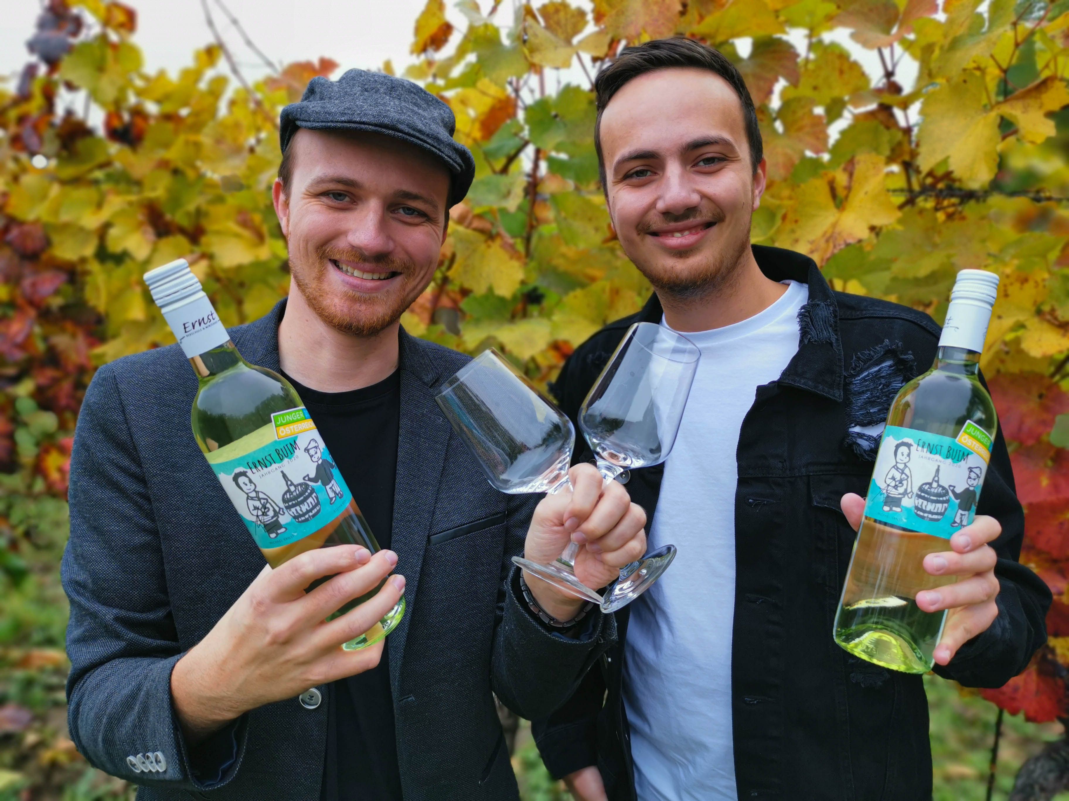 Dvaja muži držia fľaše s vínom a poháre pred jesennými vinicami.